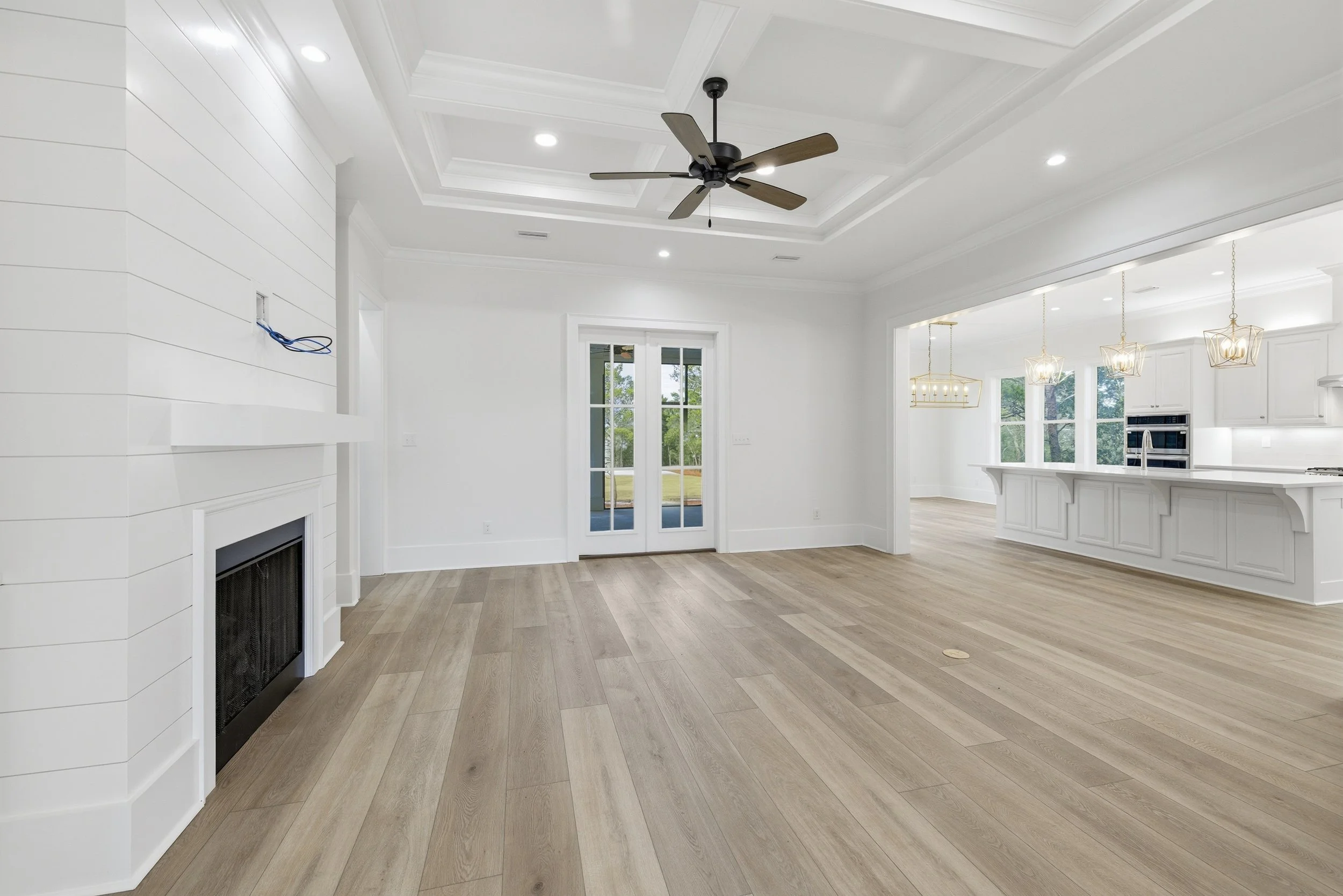 Bright, white living room with wood flooring, ceiling fan, and recessed lighting, opening to a kitchen with pendant lights and large windows.