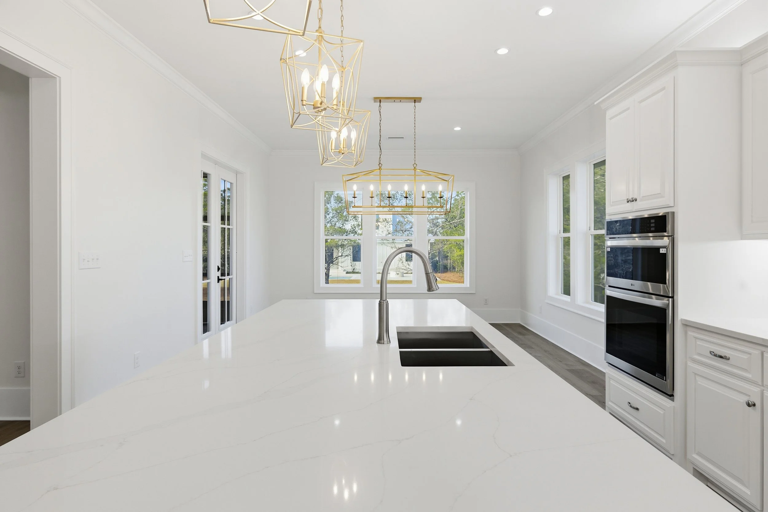 Modern kitchen with white cabinetry, a large white island with a sink, and two gold and brass light fixtures hanging from the ceiling. There are large windows and a glass door providing natural light.