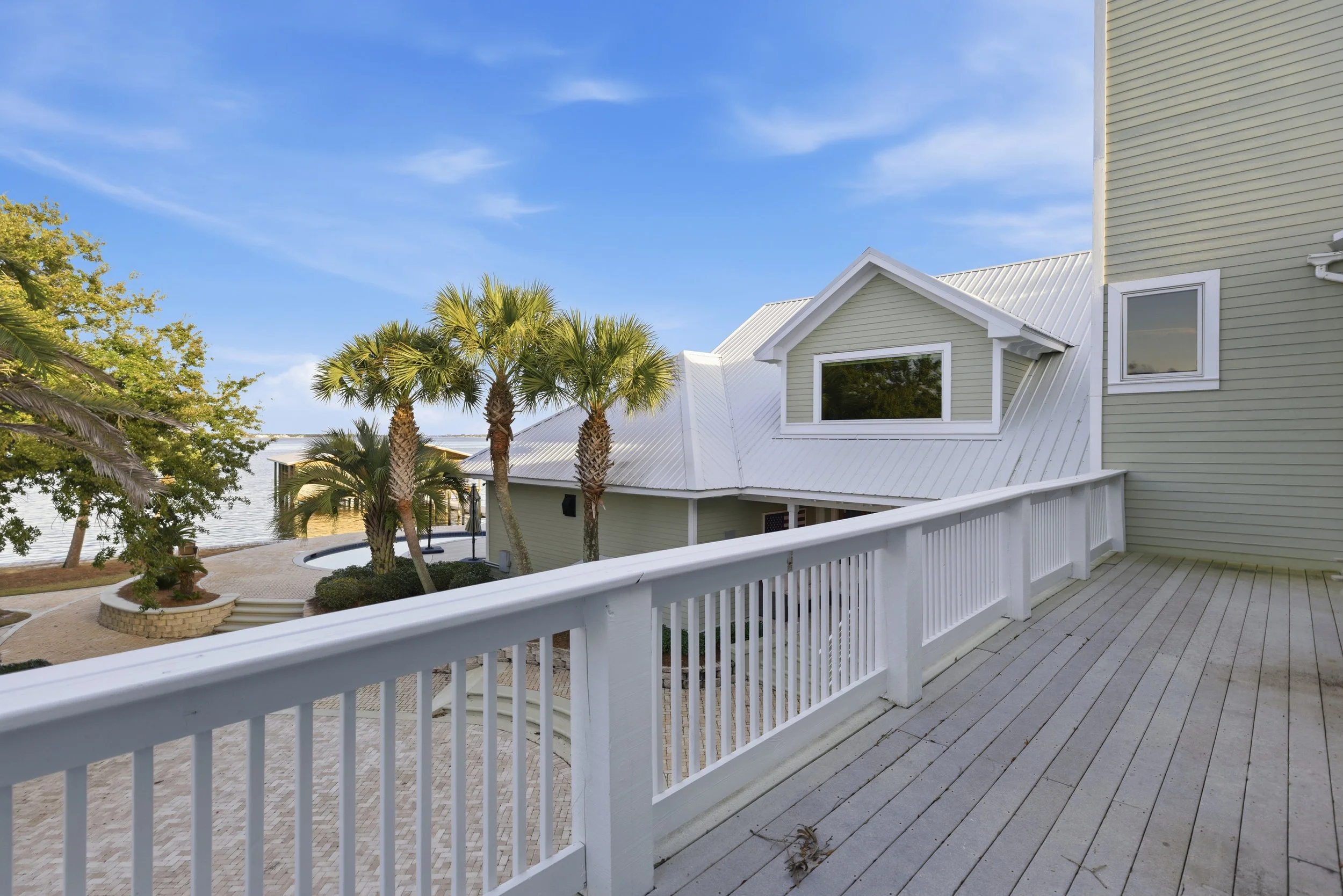 View from a balcony overlooking a backyard with palm trees, a hot tub, and a waterfront, with a house and blue sky in the background.