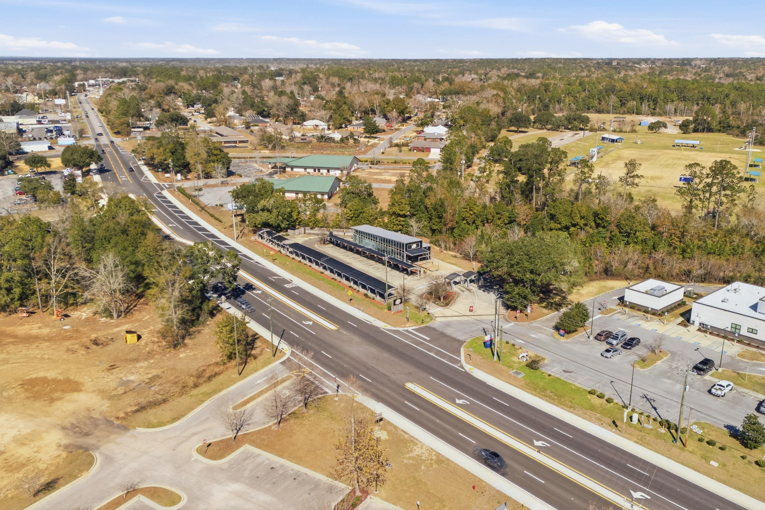 Aerial view of a suburban area with roads, trees, and buildings, including parking lots and a small shopping center, under a partly cloudy sky.
