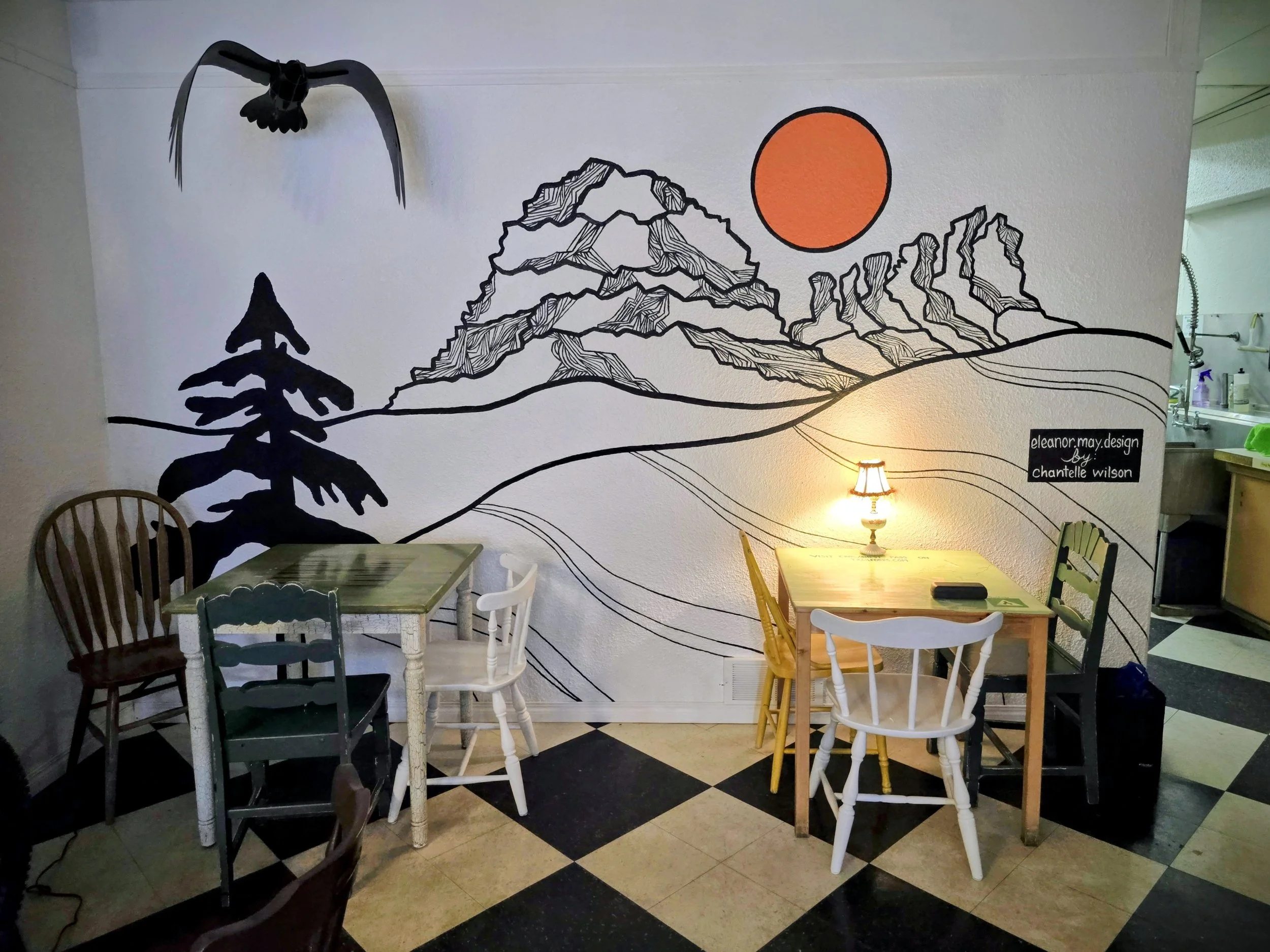 Interior of a cafe with a mural of mountain landscape and sun on the wall, four chairs, two small tables, one with a small lamp, and a chalkboard with the designer's name.