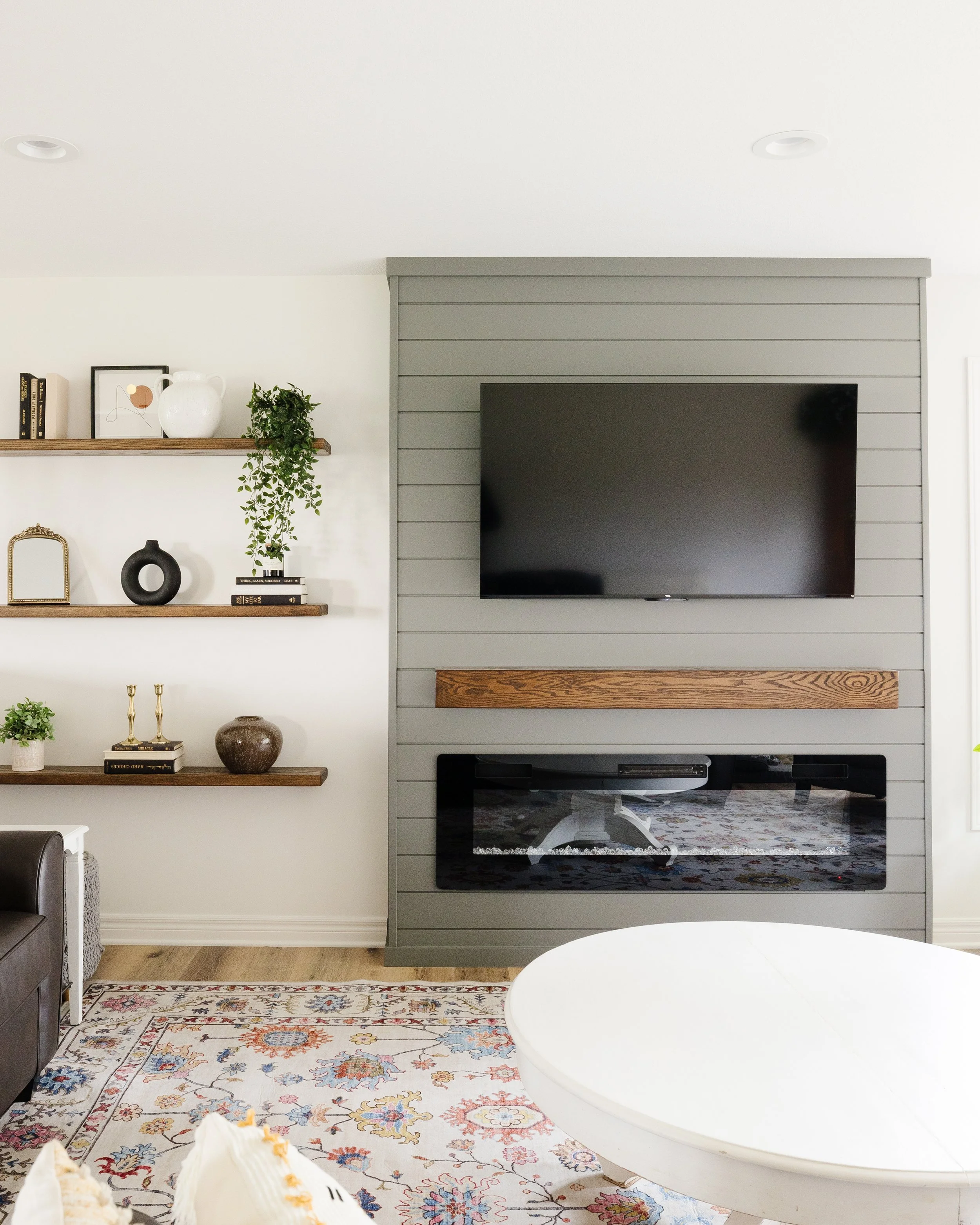 Living room with a wall-mounted TV on a gray shiplap wall, a fireplace below, and decorative shelves with books, art, and plants on a white wall to the left. There is a patterned rug, a white round table, and a brown leather chair in the room.