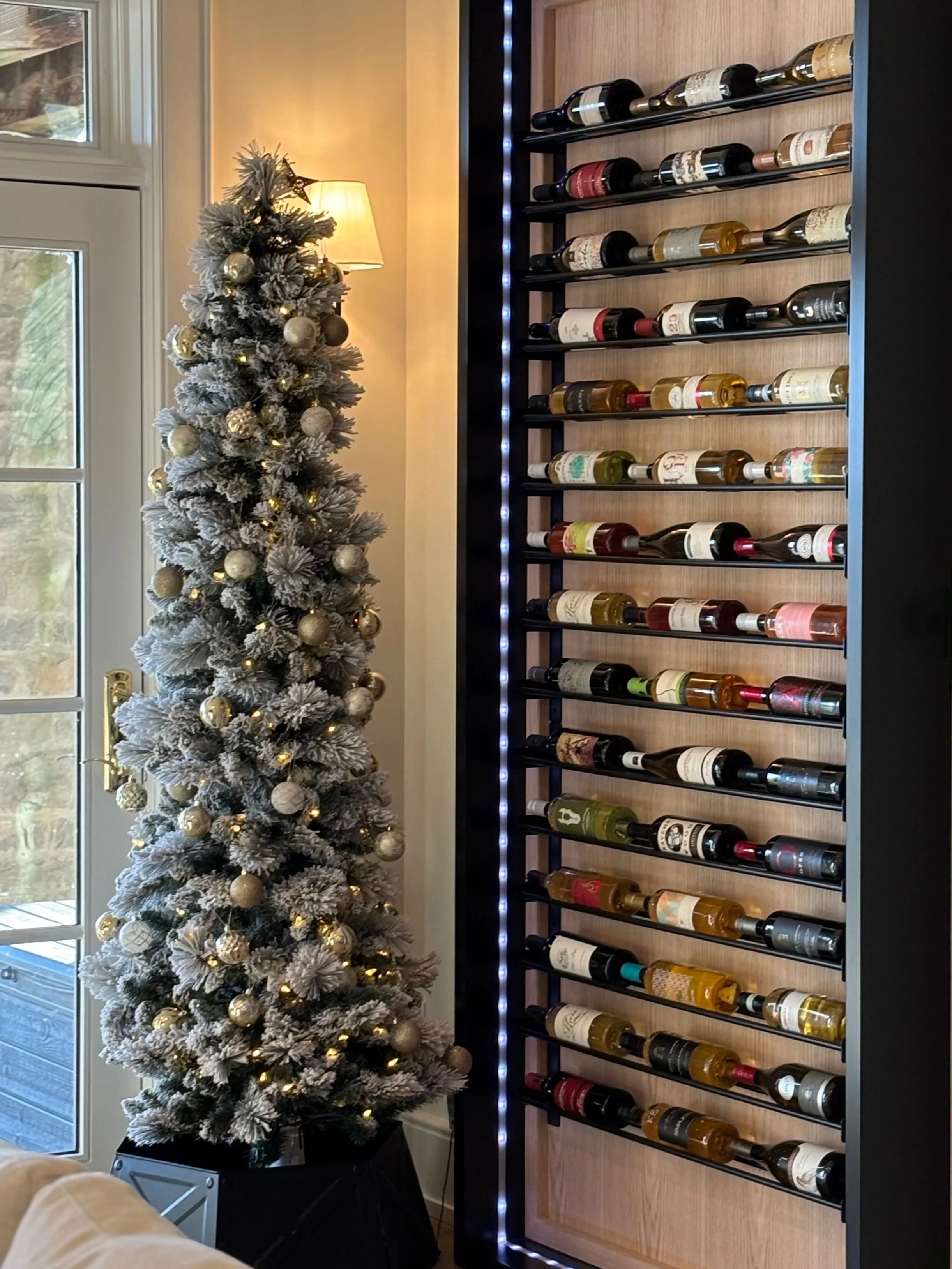 A decorated Christmas tree with ornaments and lights next to a wall-mounted wine rack filled with various bottles of wine, near a window and a lamp.