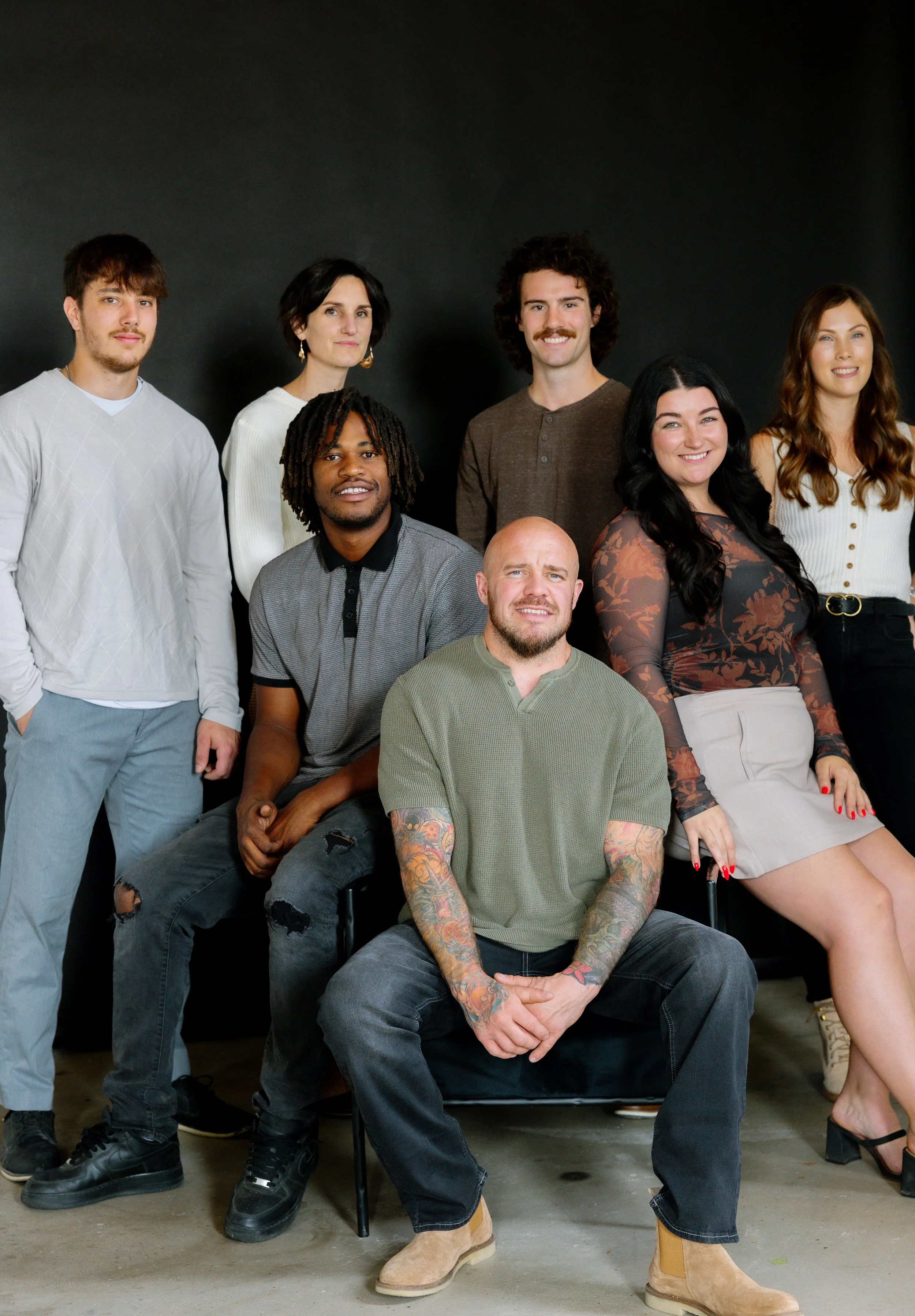 Group portrait of seven diverse young adults, some seated and some standing, against a dark background, smiling and posing for the photo.