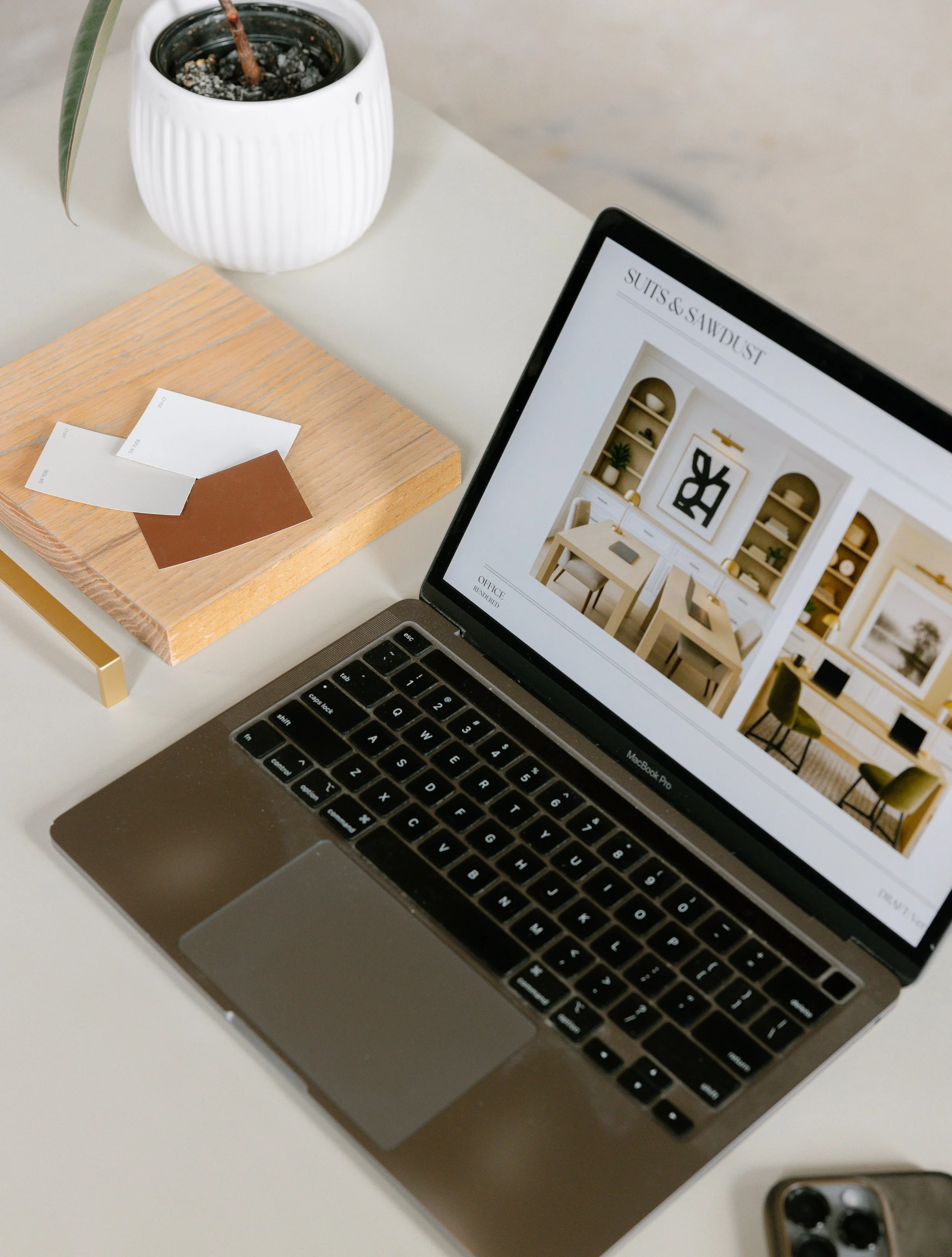 A laptop on a white desk displaying interior design images, a small wooden board with paint color samples, a white potted cactus plant, and a smartphone with a camera lens visible.