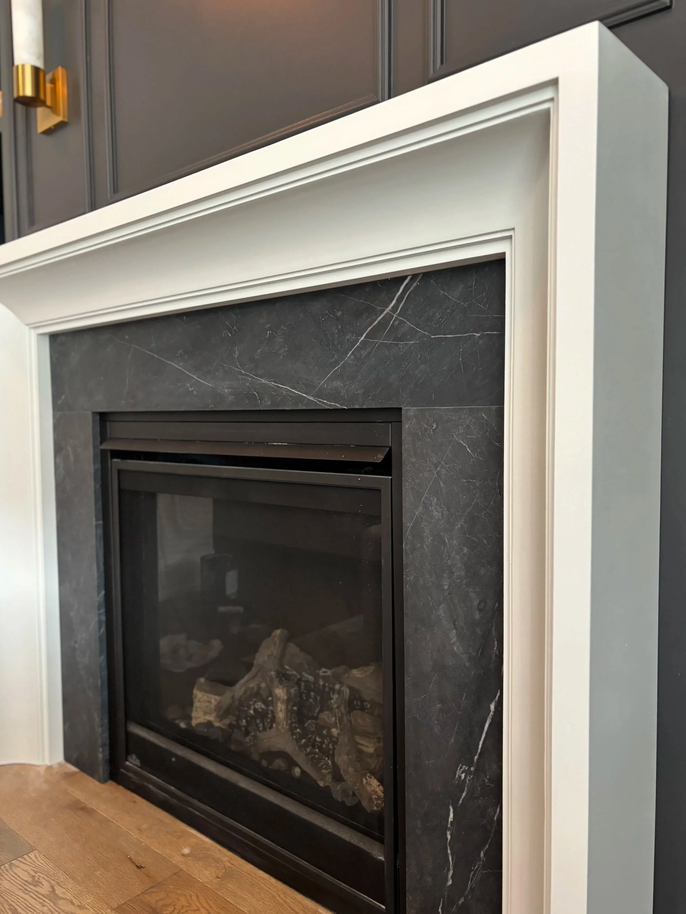 Close-up of a modern fireplace with a black stone surround, white trim, and wood logs inside. Wooden flooring is visible at the bottom.
