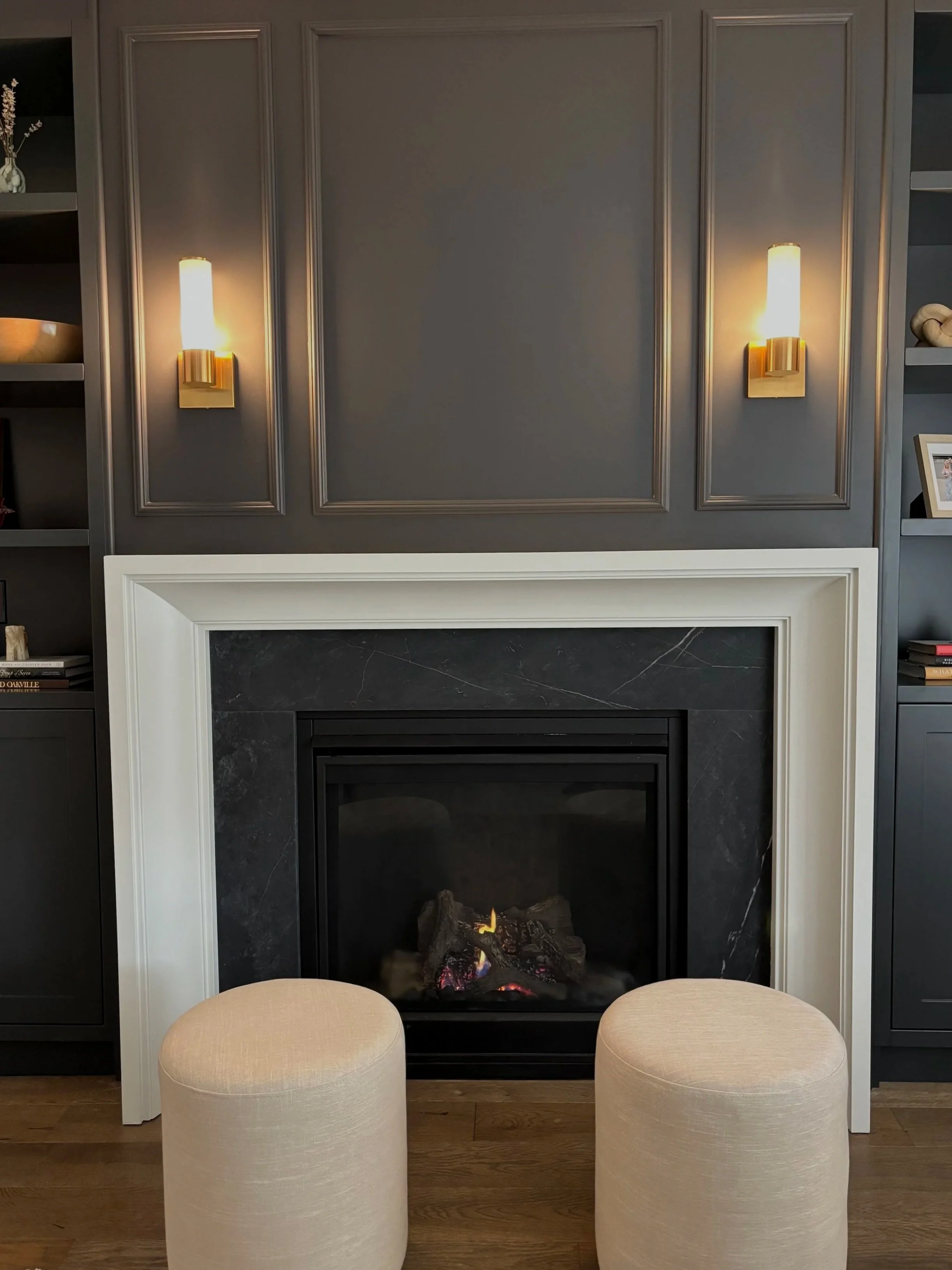 A modern living room fireplace with a black marble surround, white mantel, and a fire burning inside. Two beige ottomans are positioned in front, with gray built-in shelves on either side and two wall sconces above, providing warm lighting.