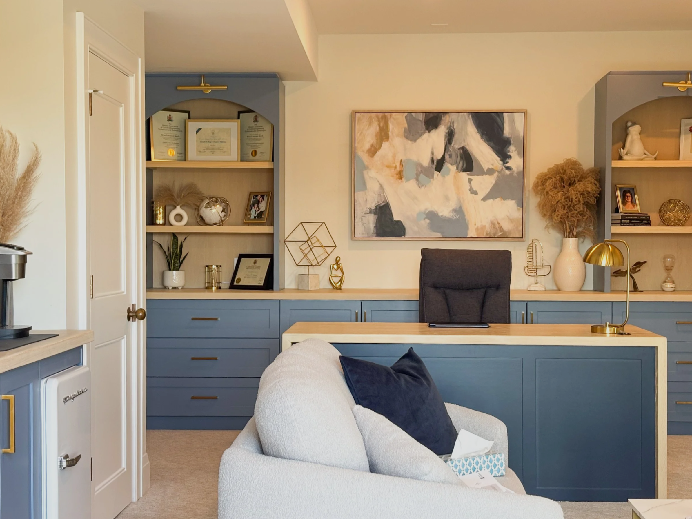 An office with a blue desk, bookshelf, and white sofa with a navy pillow.