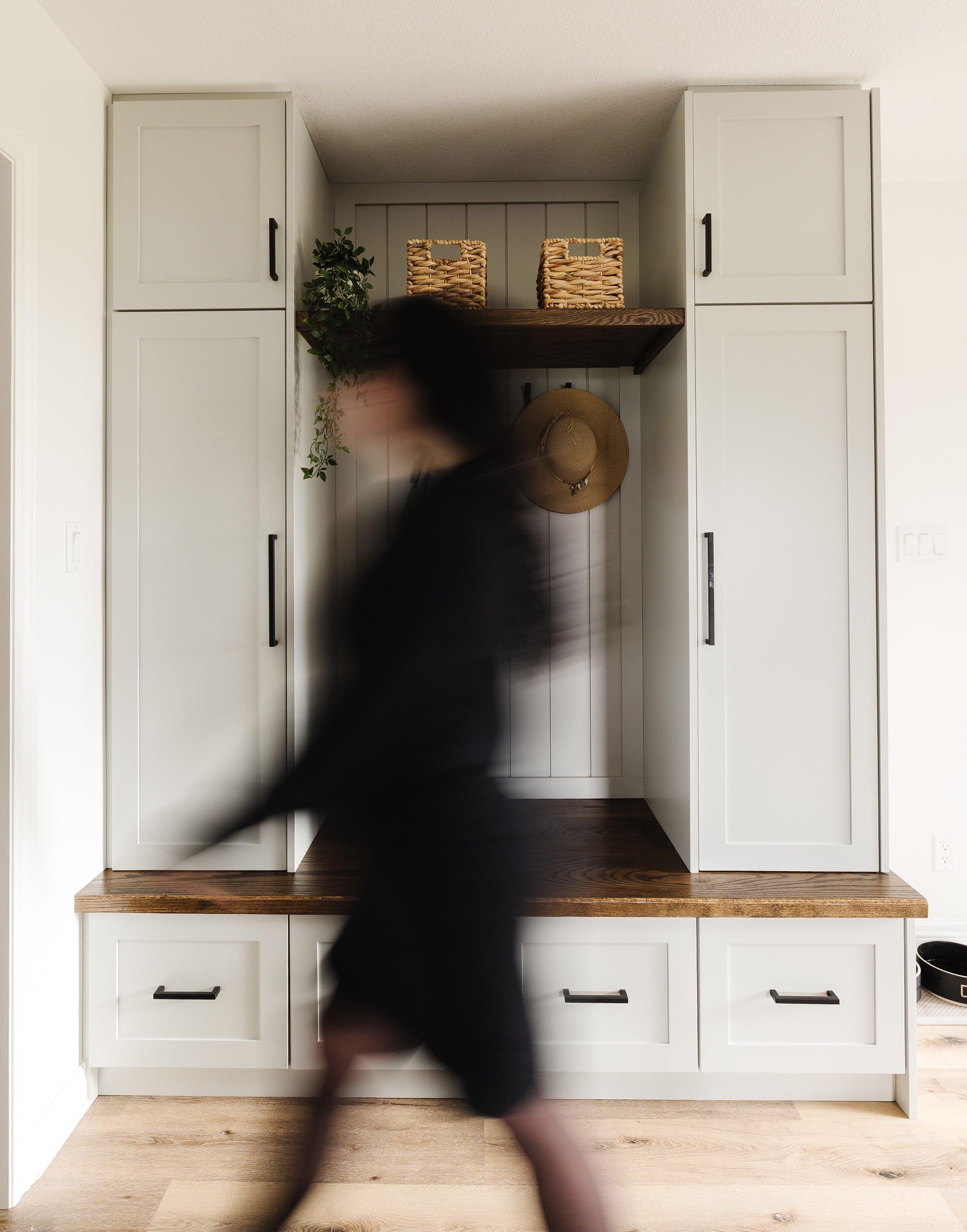 A blurred person walking past a modern white built-in cabinet with wooden accents and lower drawers in a home interior.