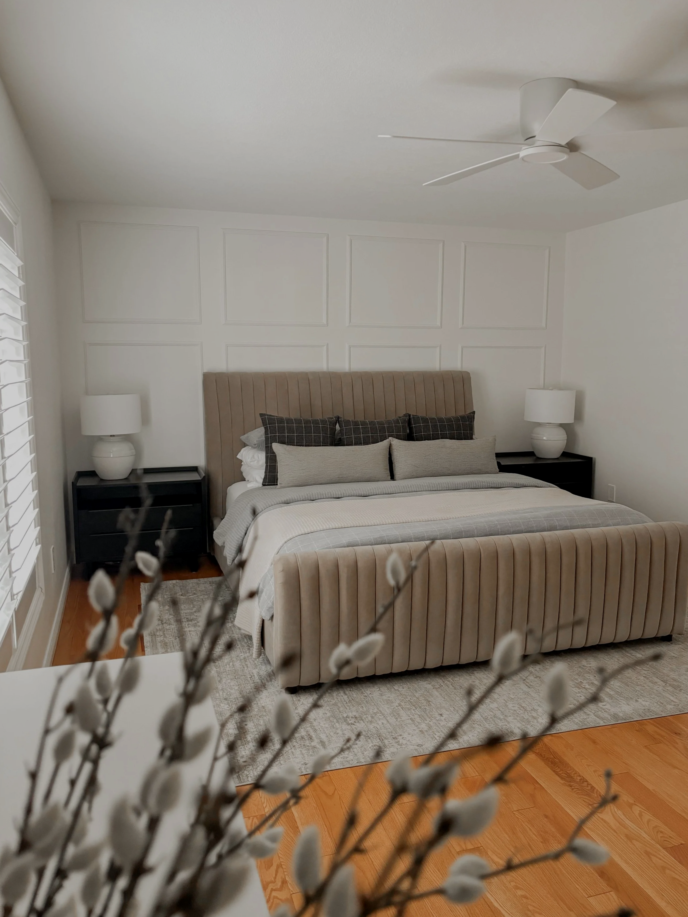 A bedroom with a beige upholstered bed, black nightstands with white lamps, and white paneled walls. A ceiling fan and a window with white shutters are visible, with blurred pussy willow branches in the foreground.