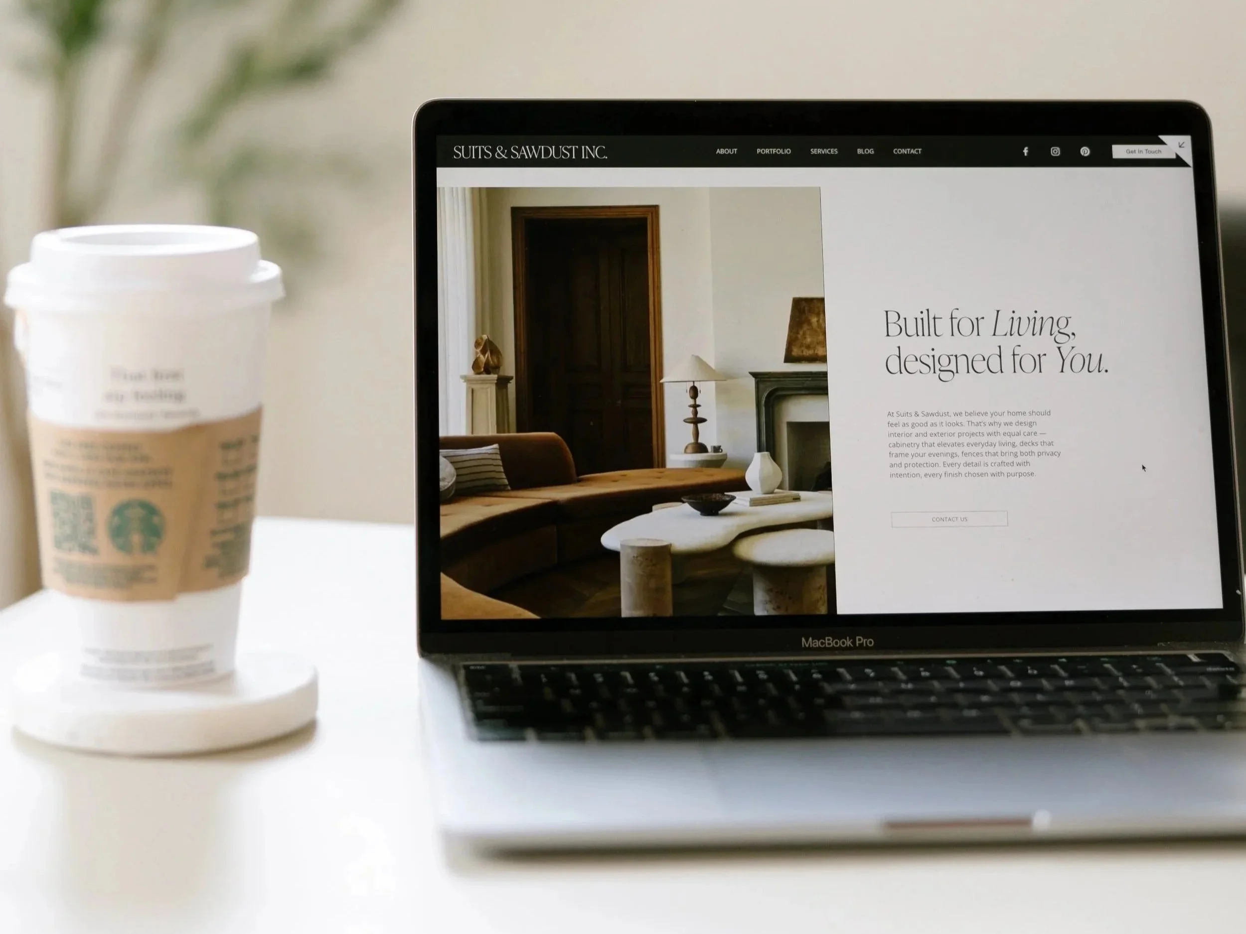 A MacBook Pro laptop on a white table displaying a website about home design, with a Starbucks coffee cup on a cardboard and foam coaster next to it.