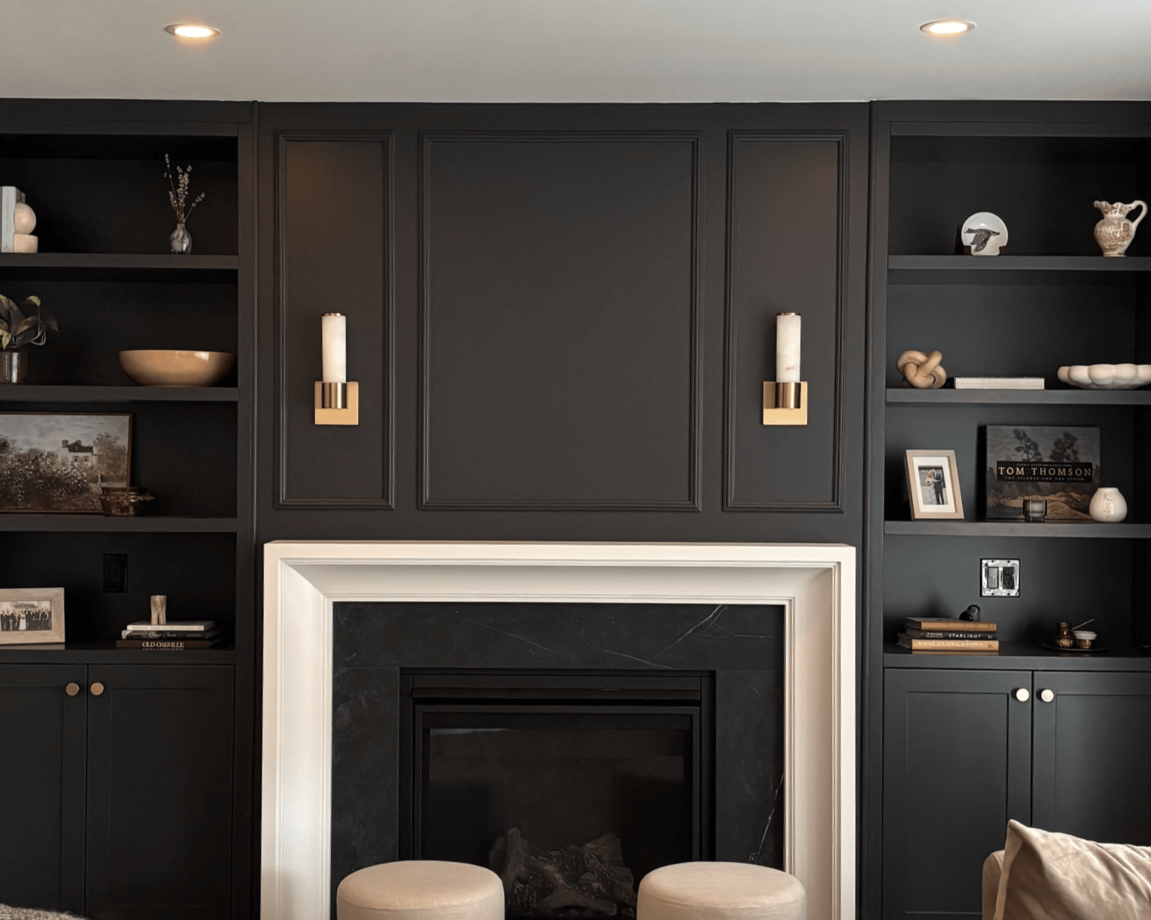 Modern living room with black built-in shelves flanking a black fireplace, decorated with various vases, books, and framed photos, illuminated by recessed ceiling lights and wall sconces.