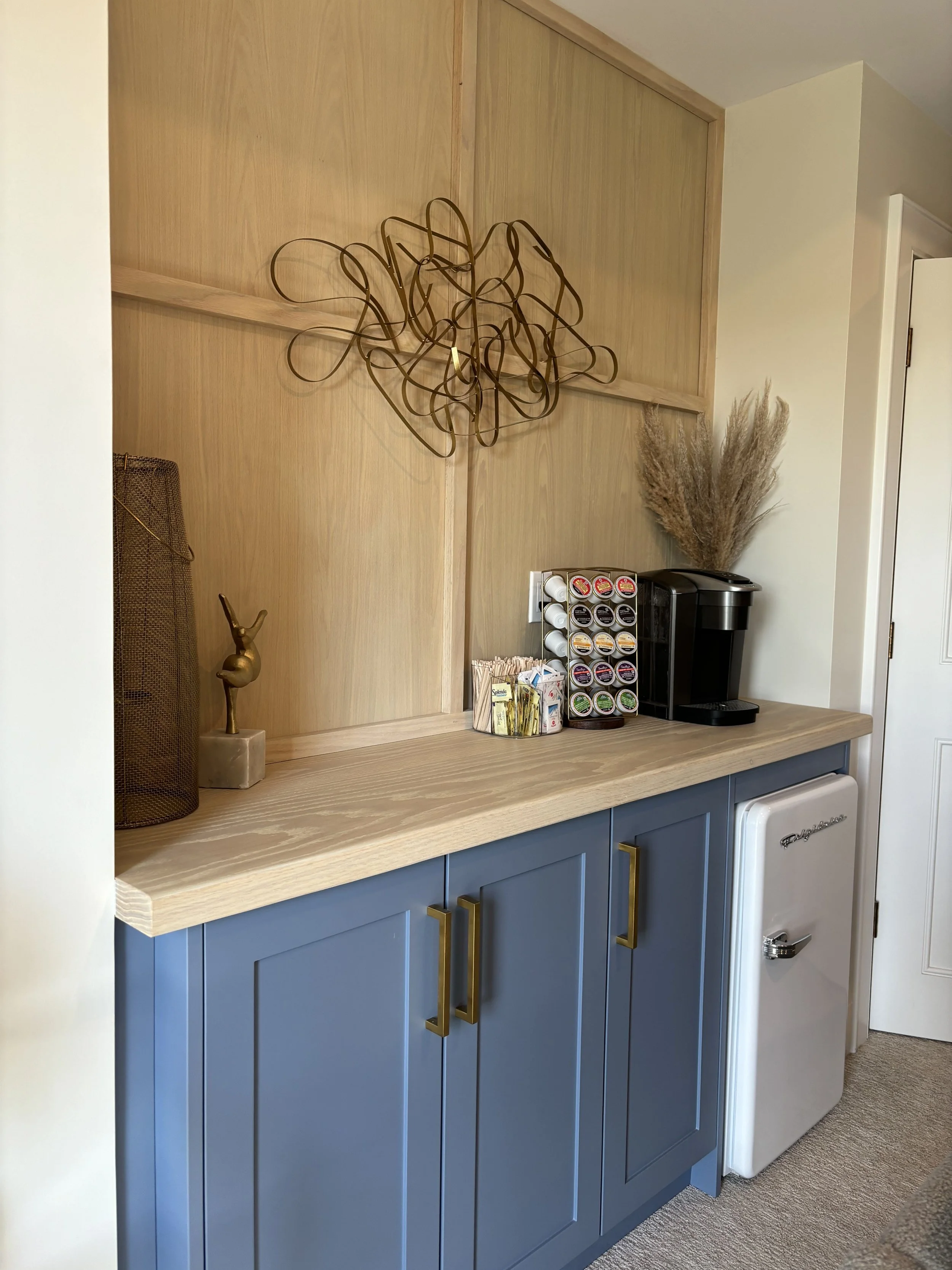 A wooden cabinet with blue doors and gold handles, a mini fridge, a black coffee machine, a coffee pod holder, and decorative dried plants and art on the wall.