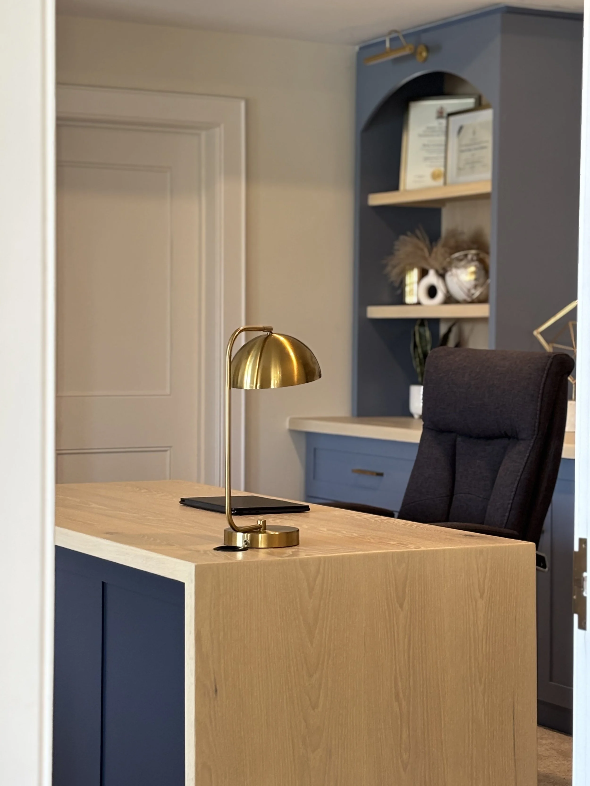 A modern office with a wooden desk, black office chair, gold desk lamp, and built-in blue bookshelf with decorative items and framed certificates.