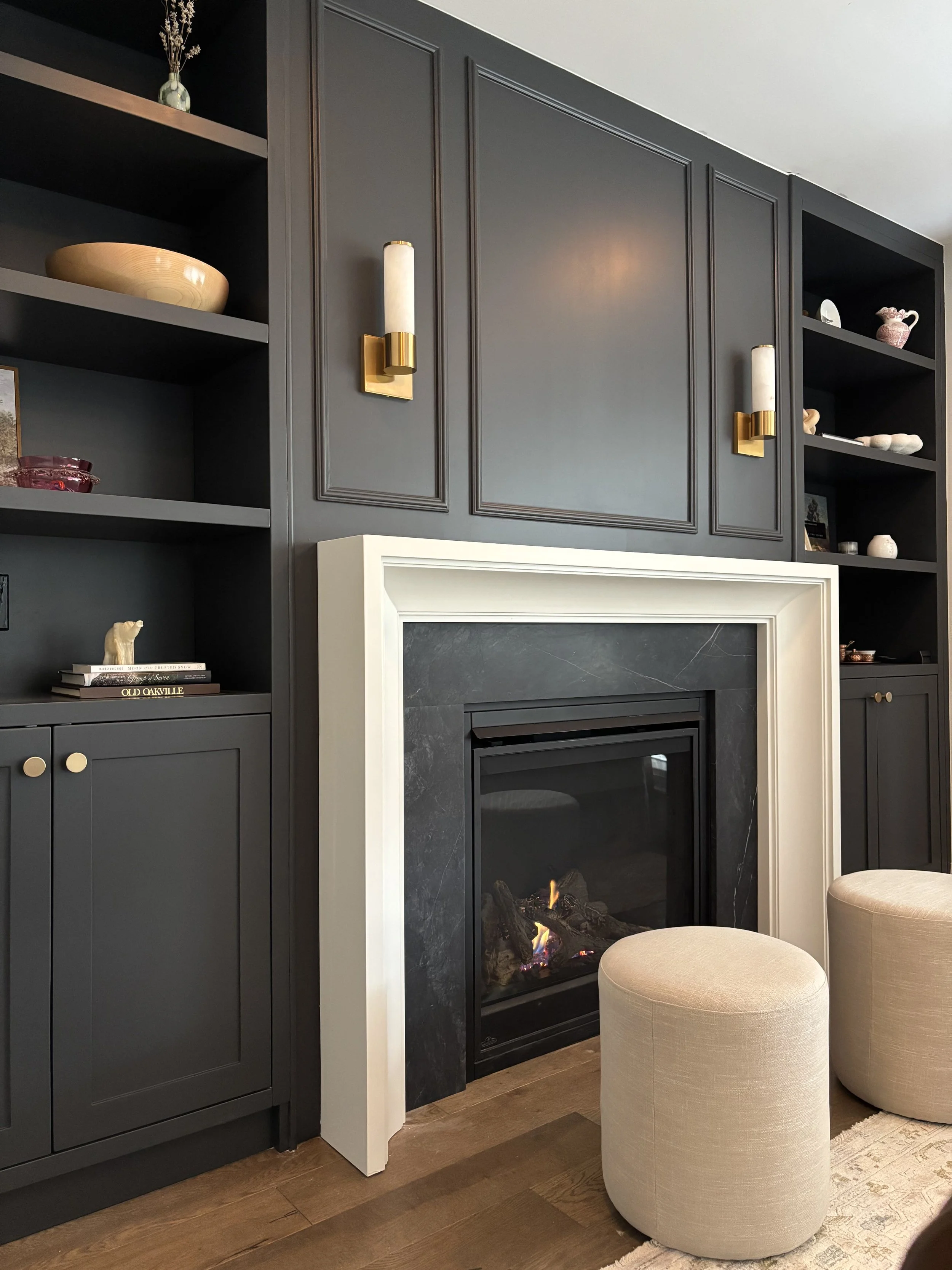 Living room corner with a black fireplace, dark gray built-in shelves and cabinets, gold wall sconces, and beige ottomans.