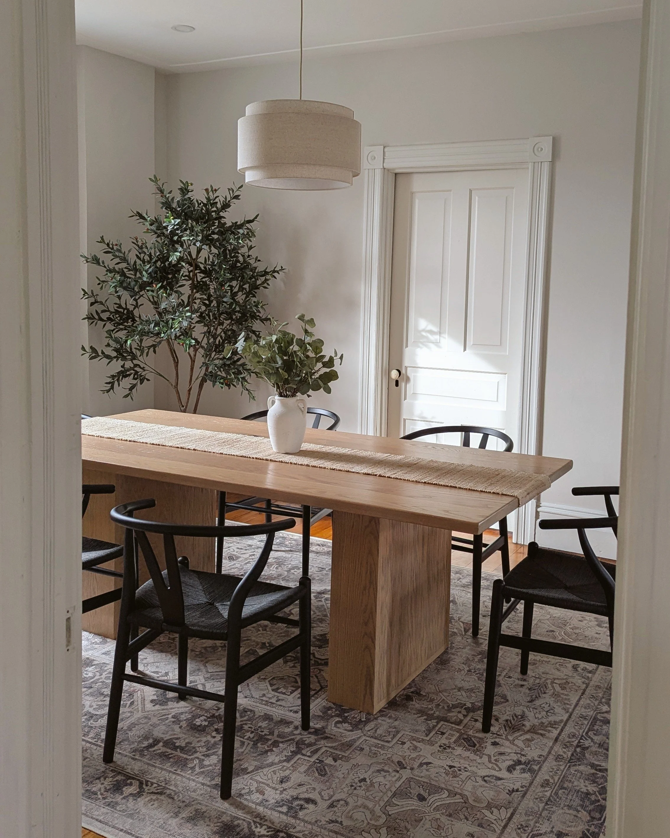 A dining room with a wooden table, four black chairs, a white vase with green foliage, a potted plant, a hanging ceiling light, and a door in the background.