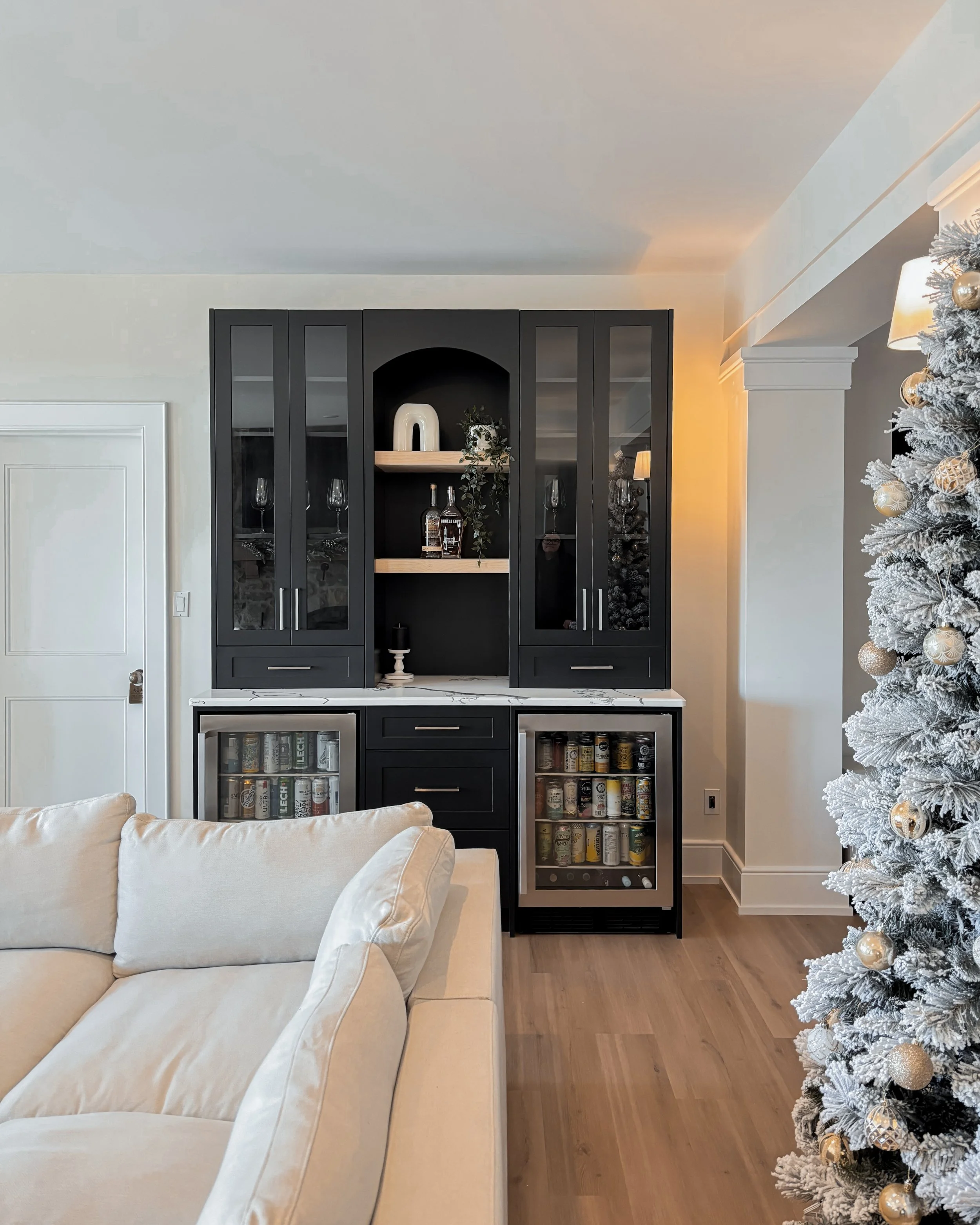 A black and white home bar with a marble countertop, glass-front cabinets, and a mini fridge filled with beverage cans, situated next to a beige sofa and a decorated Christmas tree.