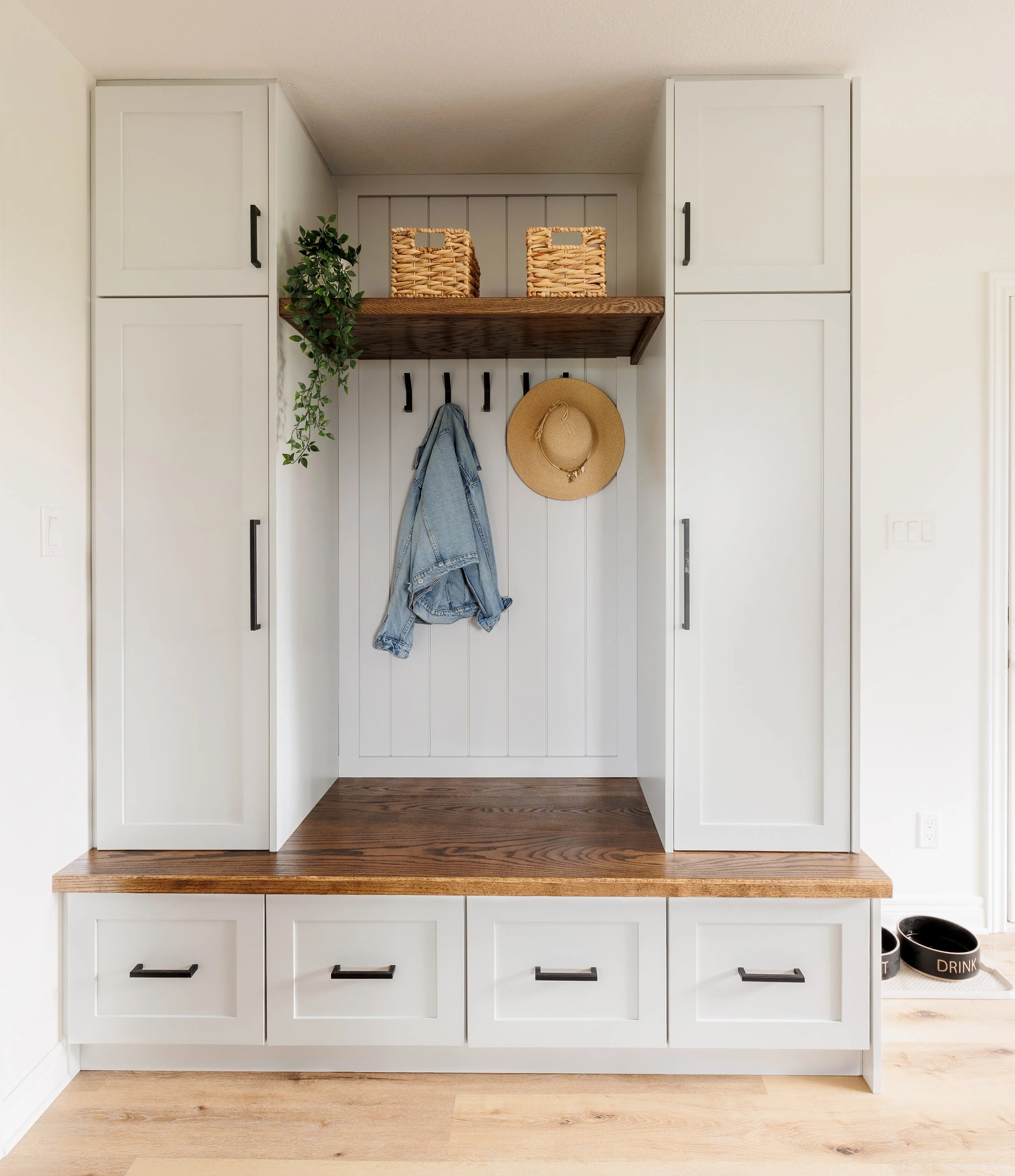 A modern entryway closet with white cabinetry, black handles, a wooden bench, and hooks on an accent wall. Items include a blue denim jacket and a straw hat. There are two woven baskets on a wooden shelf and a potted plant.