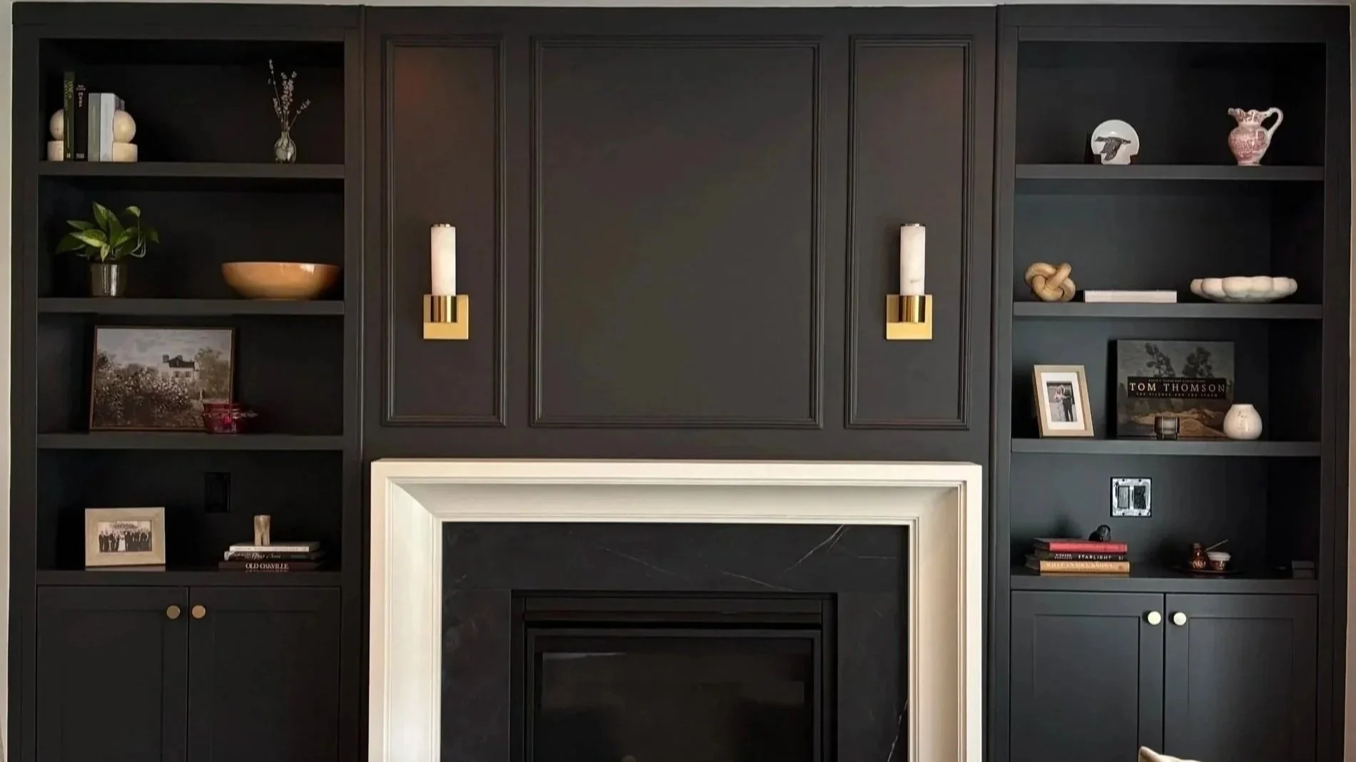 Black built-in shelves and cabinetry surrounding a fireplace, decorated with books, framed photos, small sculptures, and a potted plant.