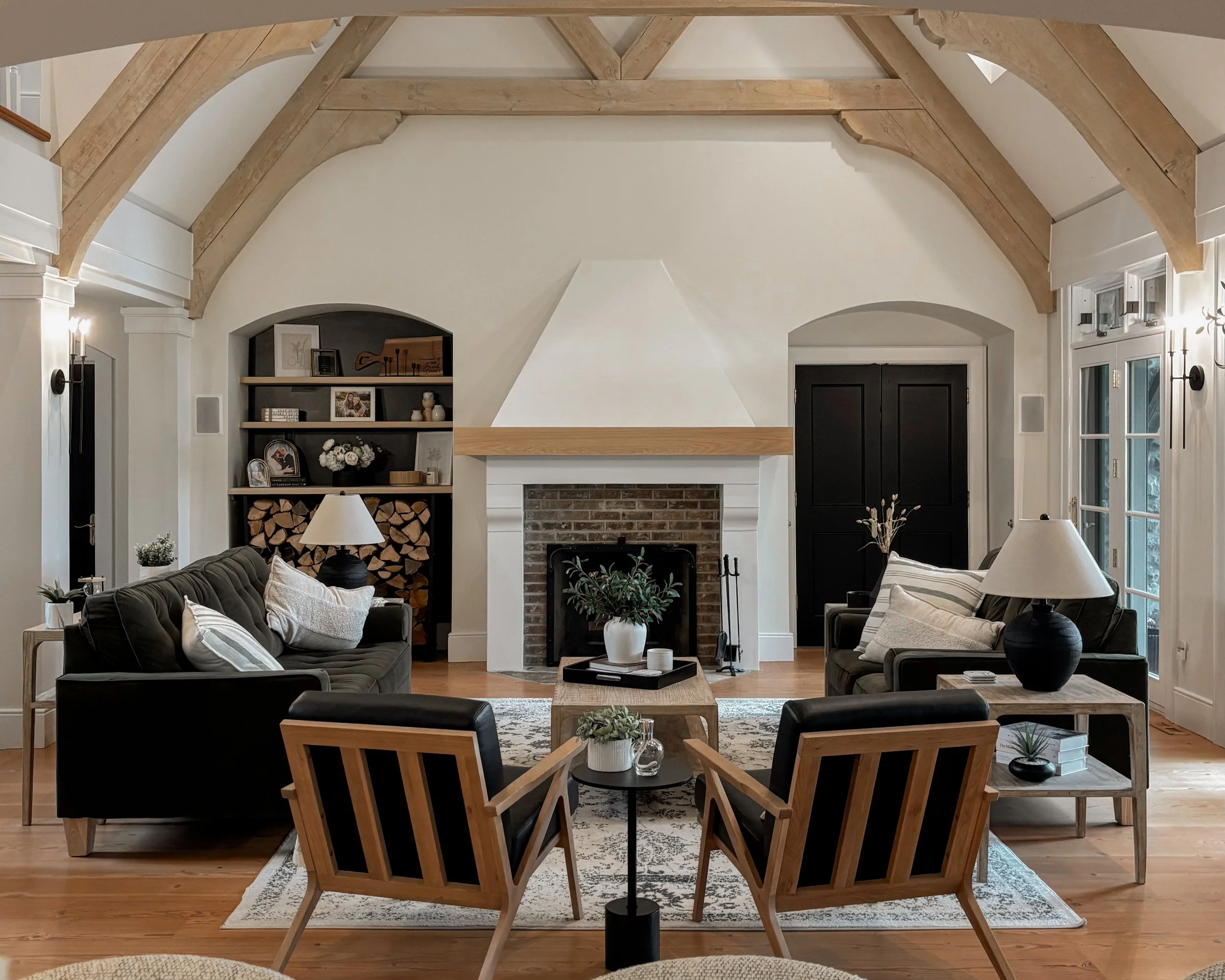 Living room with a fireplace, two black sofas with white cushions, two wooden armchairs, a coffee table with a potted plant, a bookshelf filled with firewood, and large windows.