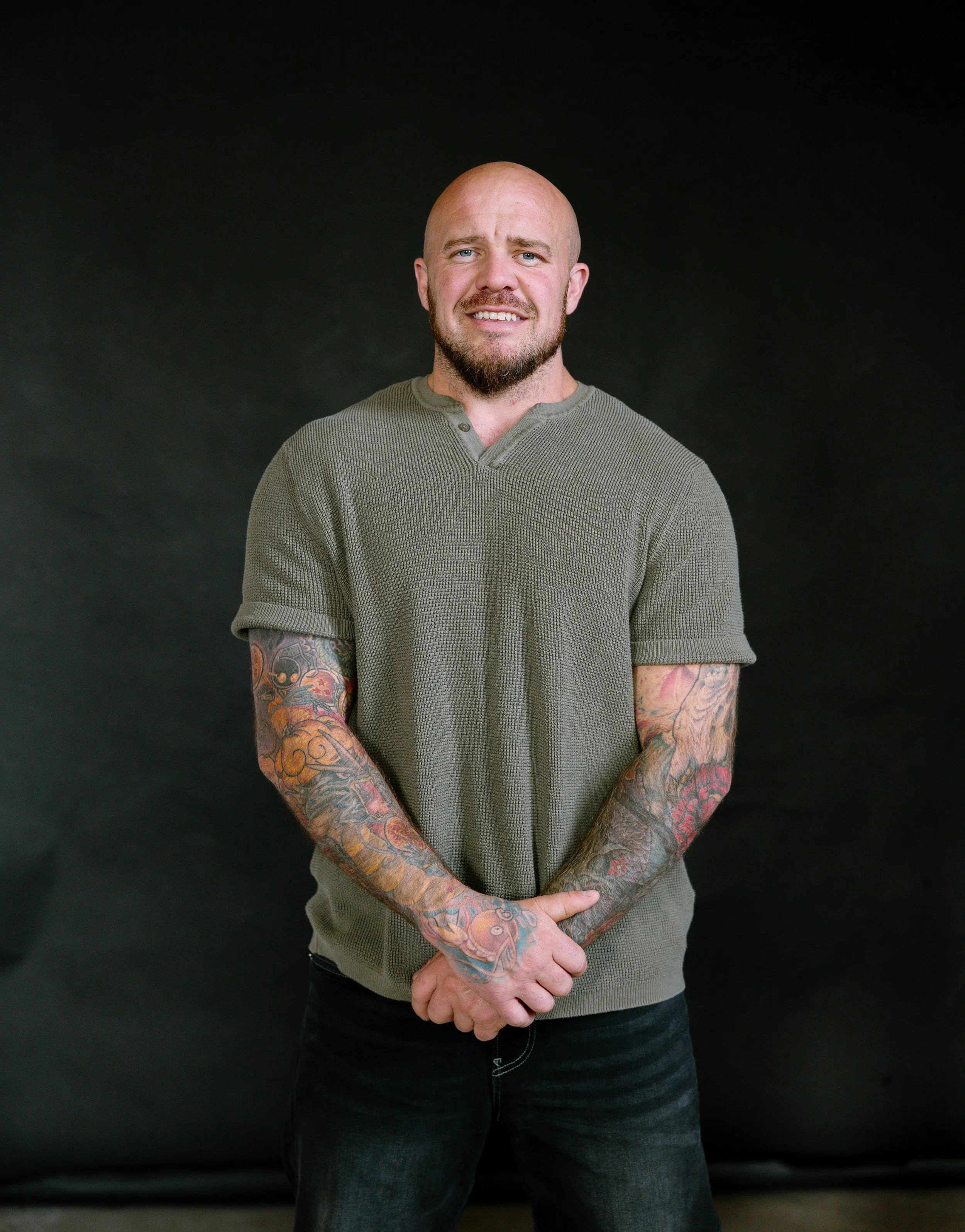 A man with a shaved head, beard, and tattoos on both arms, wearing a green short-sleeved knit shirt and black jeans, standing against a black background with a friendly expression.