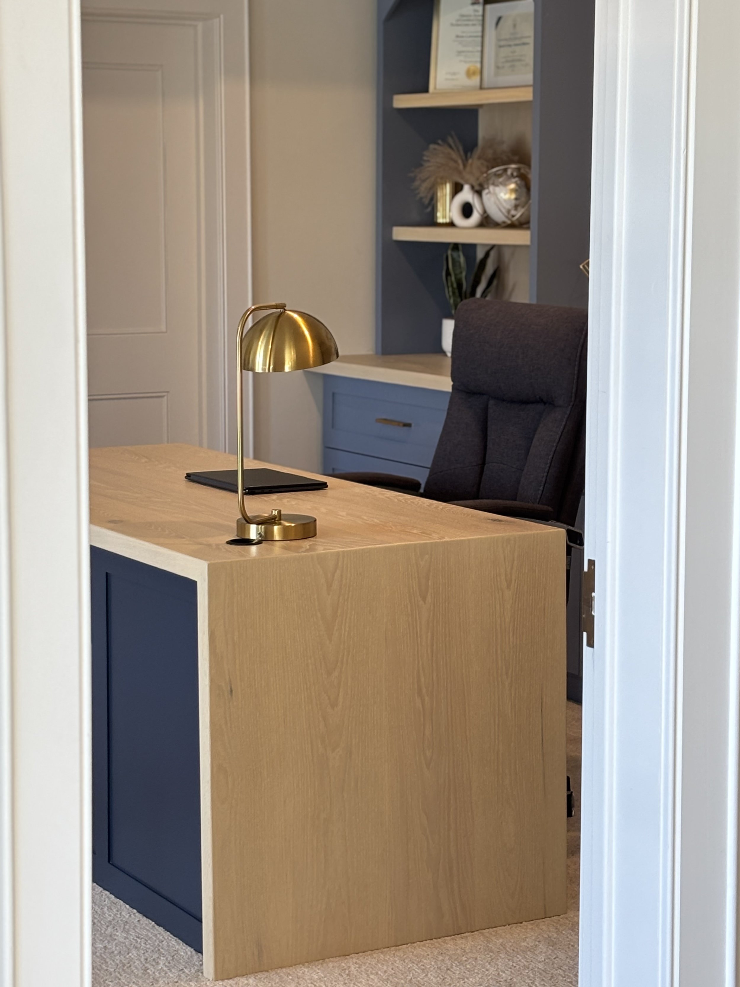 View of a modern office corner through a doorway, featuring a wooden desk with a black notebook and a gold desk lamp, a upholstered executive chair, and built-in shelving with decor and books.