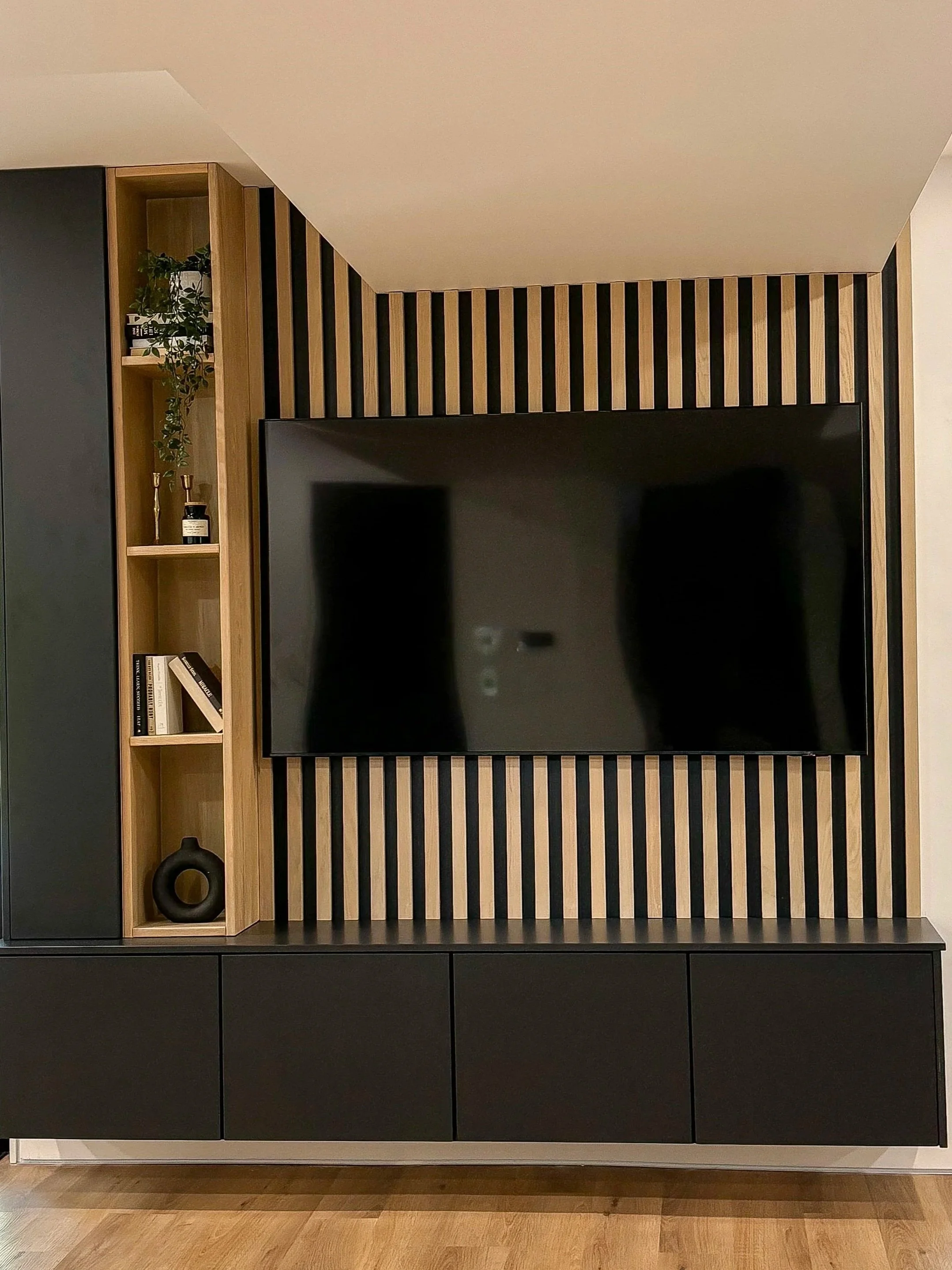 Modern living room entertainment wall with a large flat-screen TV mounted on a wooden slat panel, black cabinets below, and a vertical open shelving unit with plants, books, and decorative objects on the left.