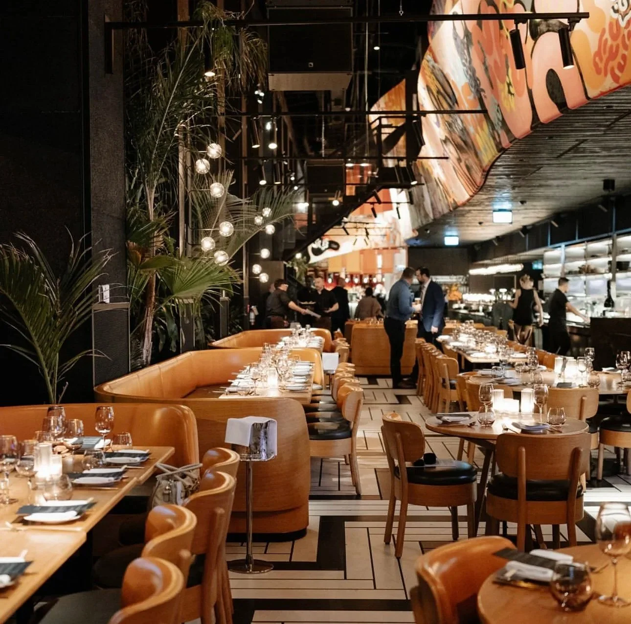 Elegant restaurant interior with wooden tables set with glasses, plates, and cutlery, warm lighting, and modern decor including hanging lights and potted plants; restaurant staff assist customers while a few guests are seated.