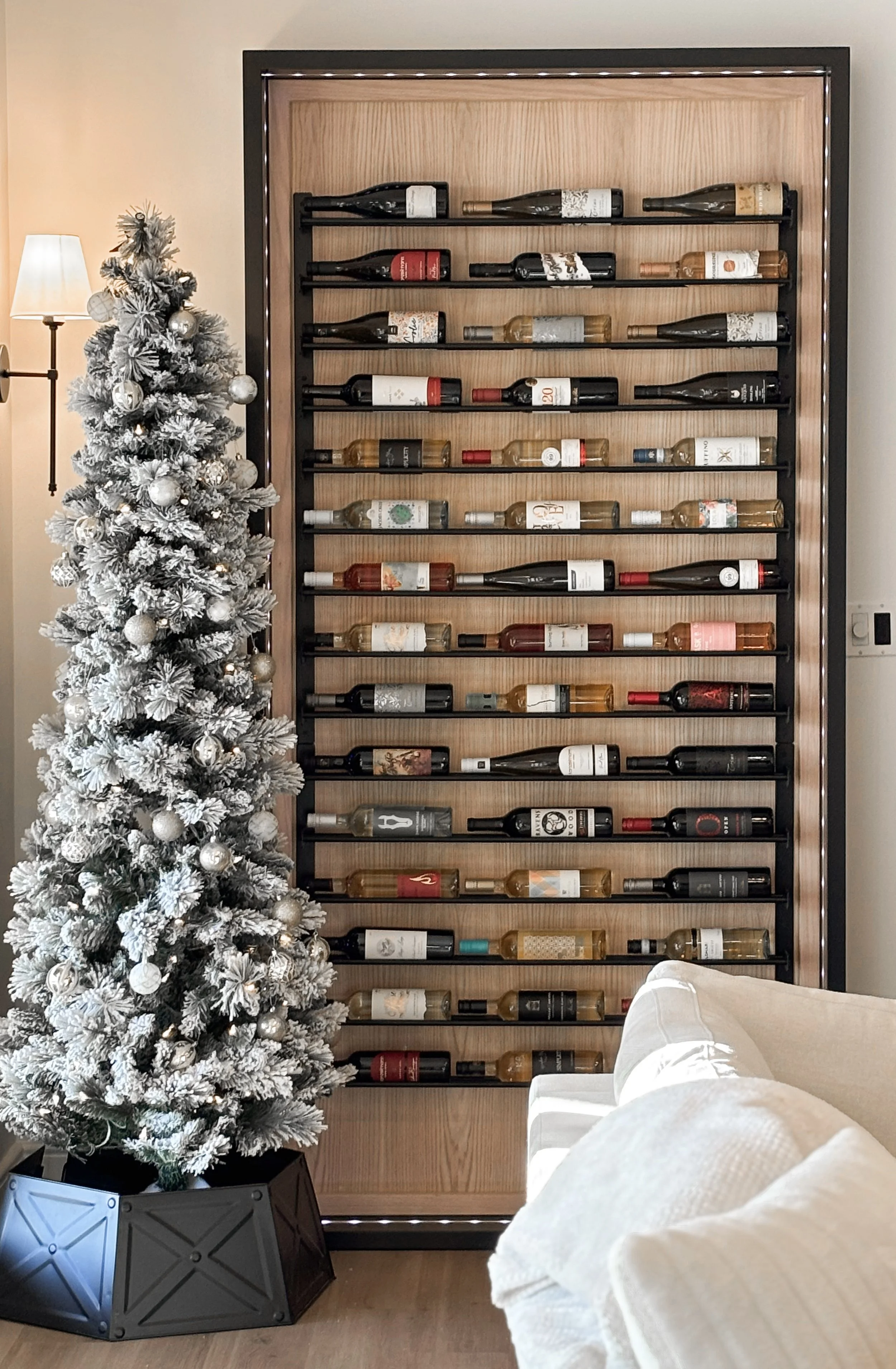 A white artificial Christmas tree decorated with silver ornaments standing next to a wine rack filled with bottles in a living room.