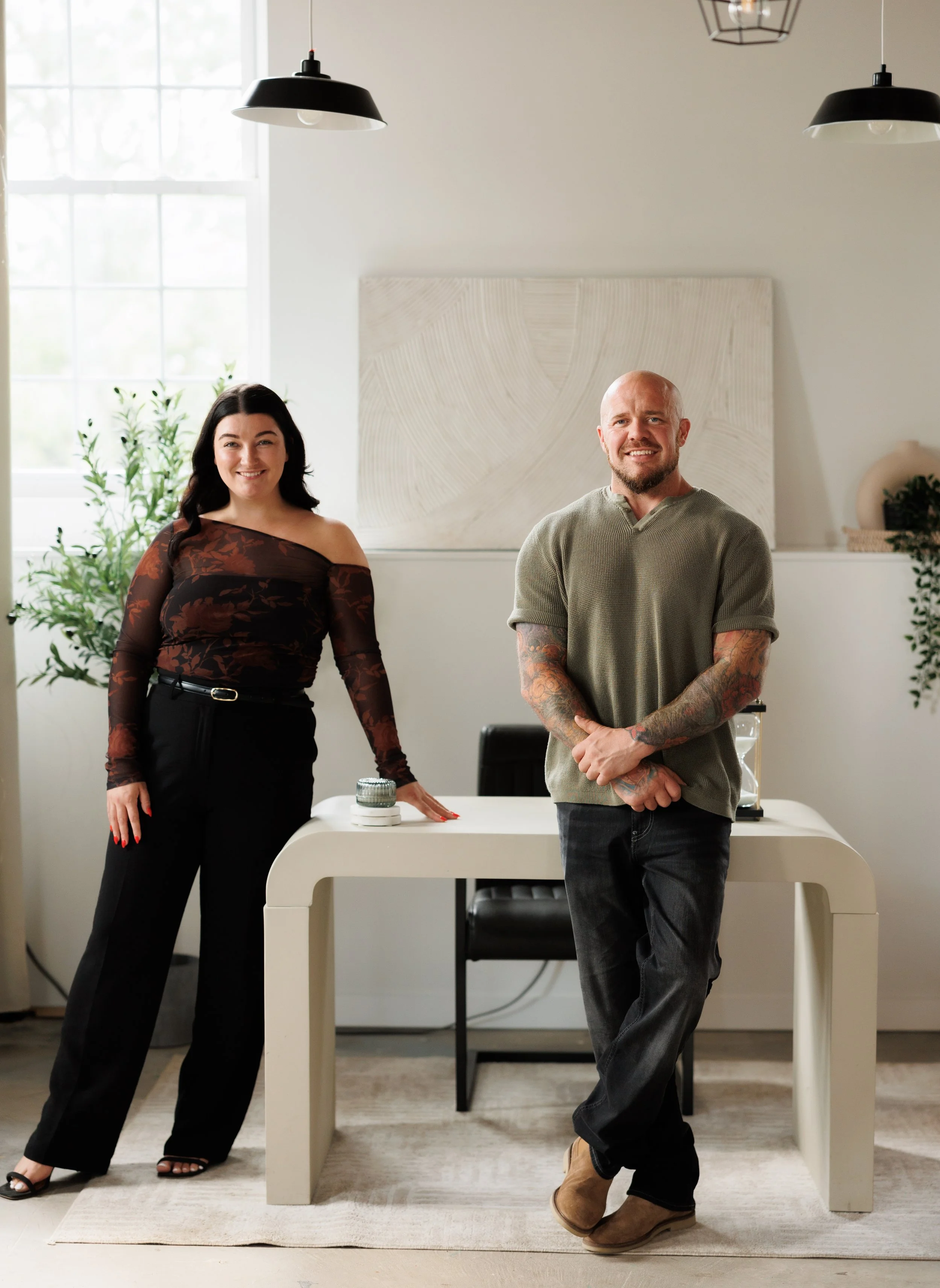 A woman and a man smiling in a bright, modern office space with potted plants, a white desk, and art on the wall.