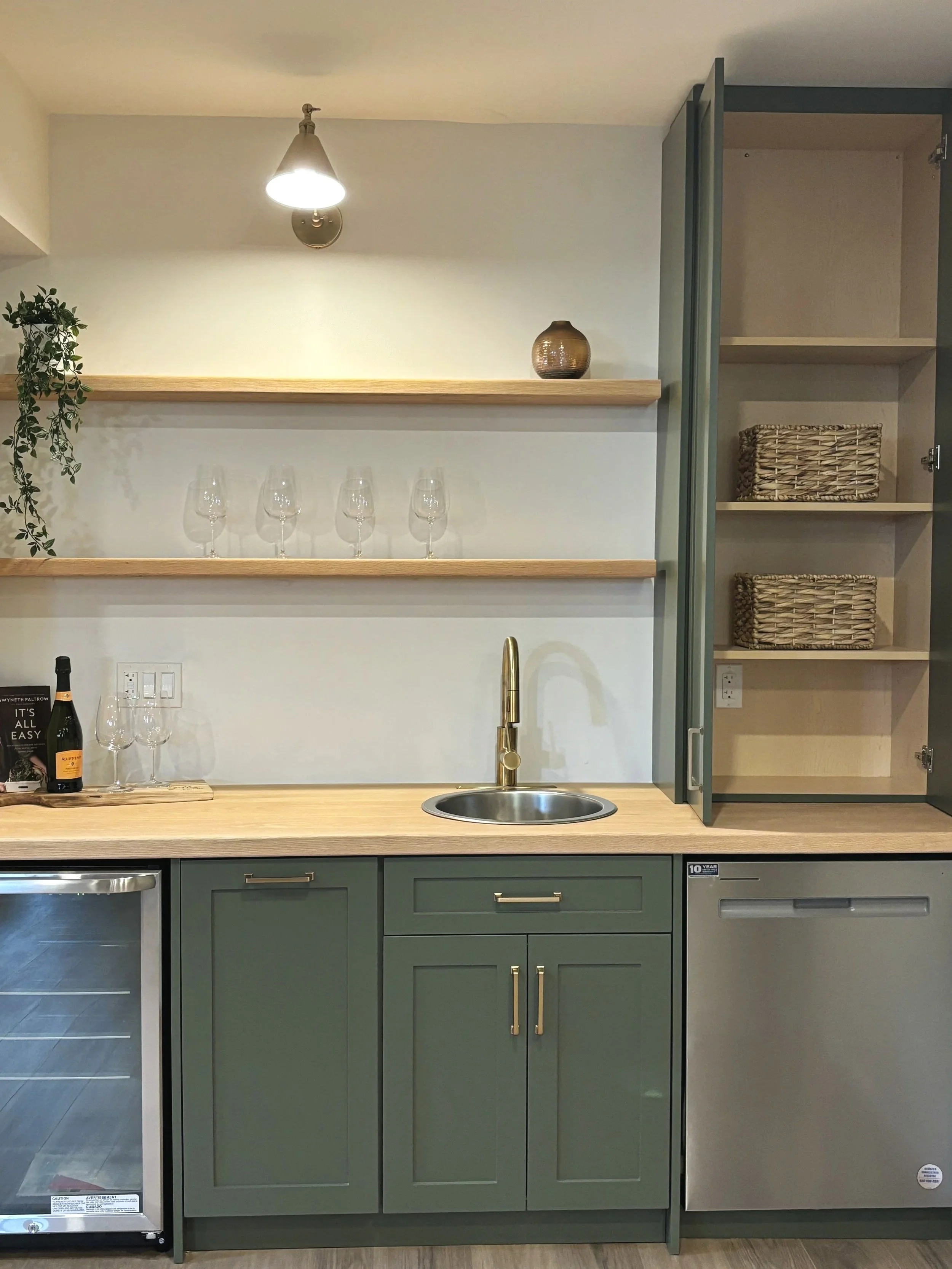 Kitchen with green cabinets, a wooden countertop, a small sink with a gold faucet, open shelves with wine glasses and decorative items, and a small refrigerator.