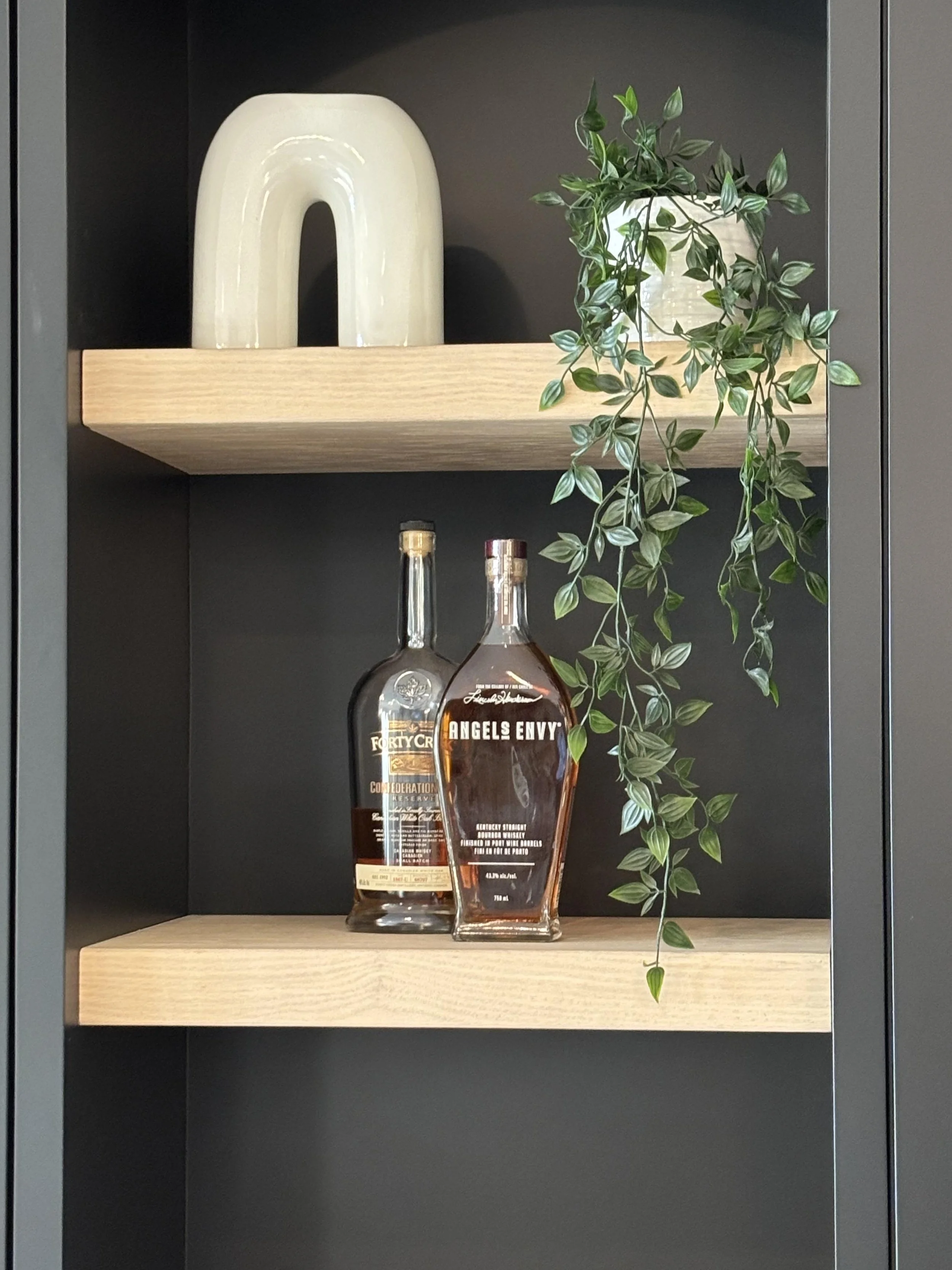 A black shelf with two wooden shelves inside. The top shelf has a white decorative vase and a green trailing plant. The bottom shelf holds two bottles of alcohol, one labeled 'Forty Cr' and the other 'Angels Envy'.