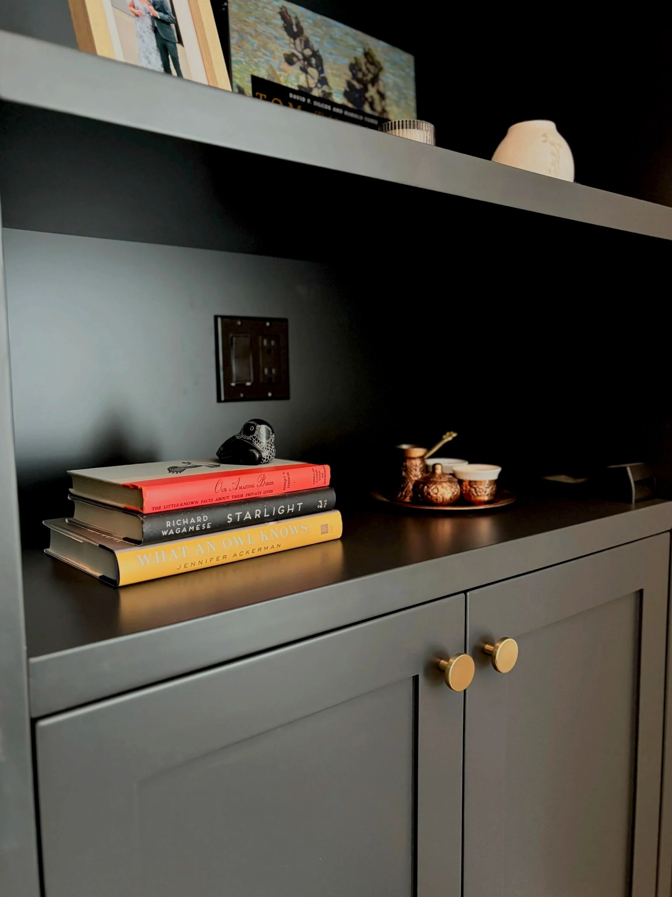 A gray cabinet with gold knobs, with a stack of three books and some decorative items on top, including a small black bird figurine and a copper-colored object with vessels. A TV is mounted above, showing a cartoon or animated scene.