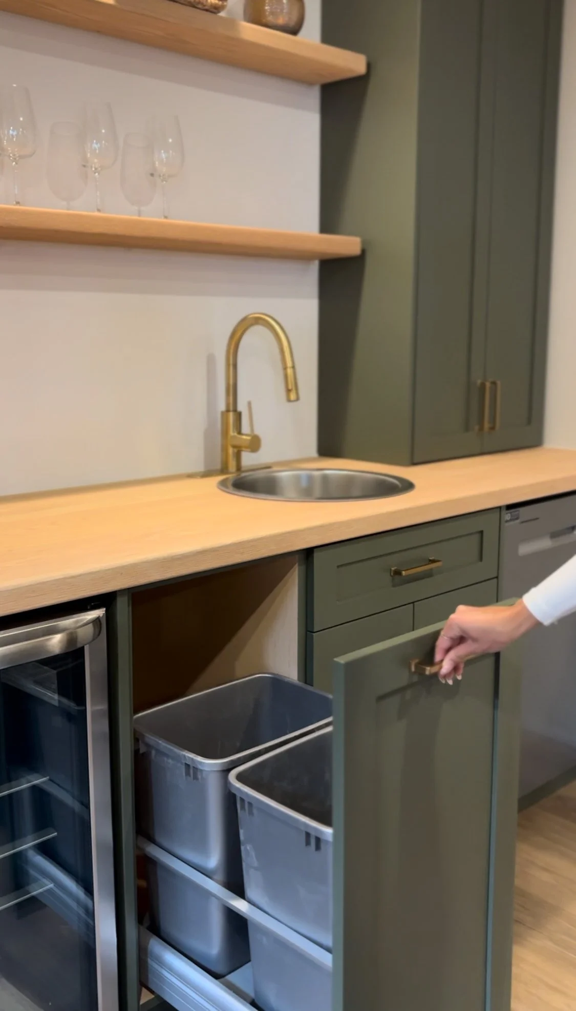 Kitchen with green cabinets, a wooden countertop, gold faucet, open cabinet drawer, shelves with glasses, and bins inside a glass-front refrigerator.