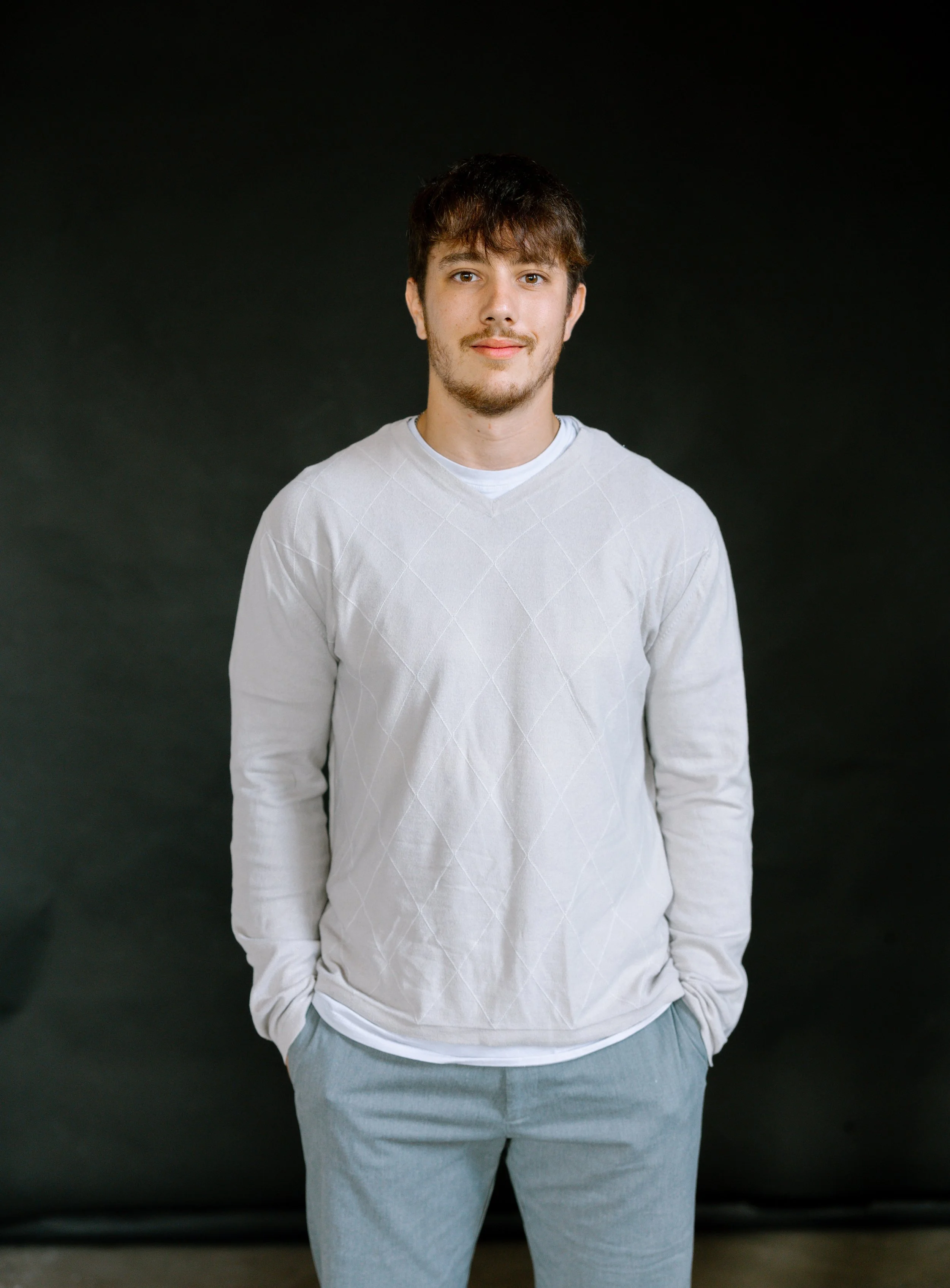 Portrait of a young man with brown hair, light beard, wearing a white sweater and light gray pants, standing against a black background.