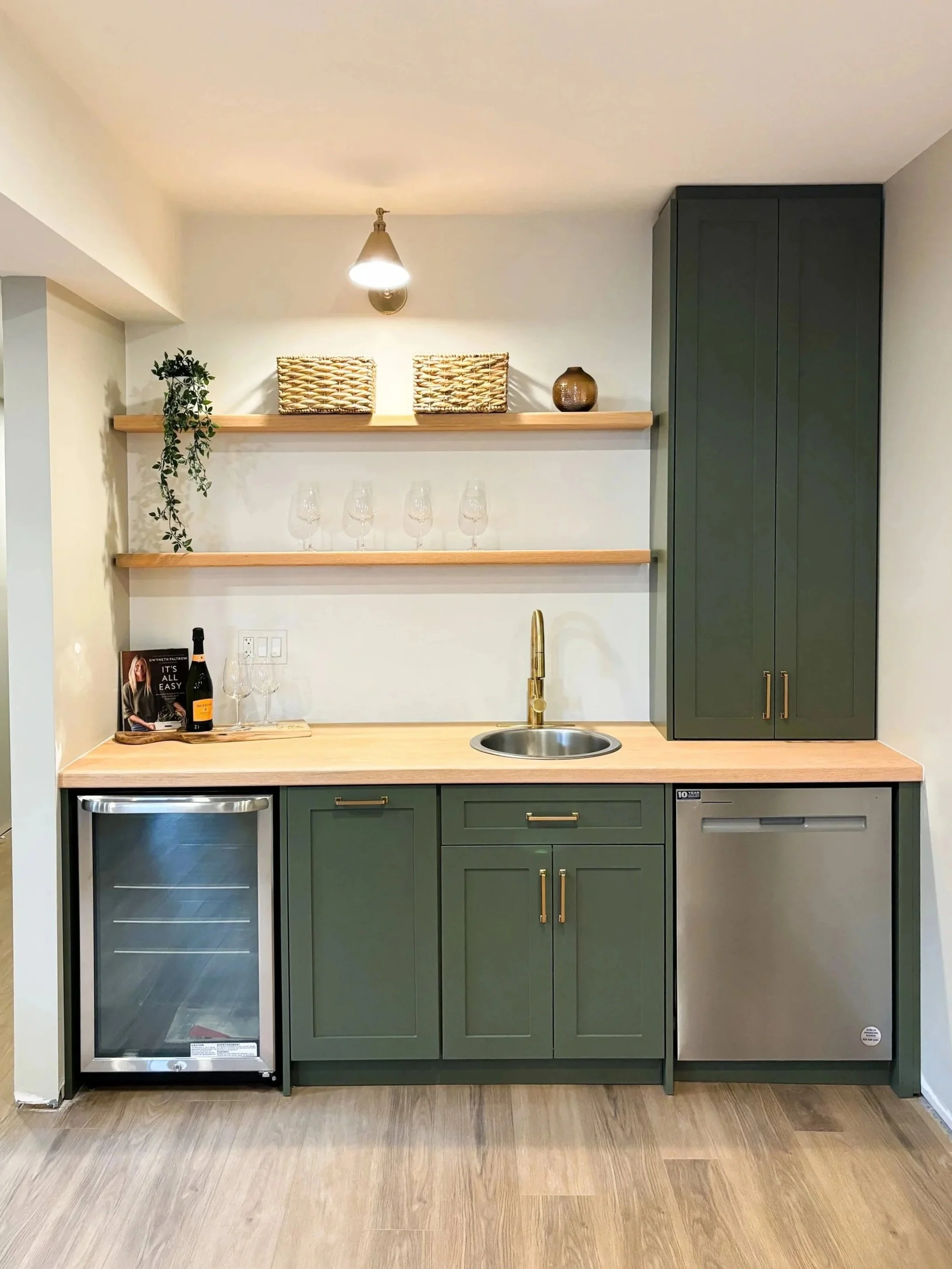 Green kitchen cabinet with a wooden countertop, small refrigerator, and mini wine fridge. Open shelving with decorative items, a wine bottle, glasses, and a potted plant. Gold faucet and cabinet handles, with a small sink.