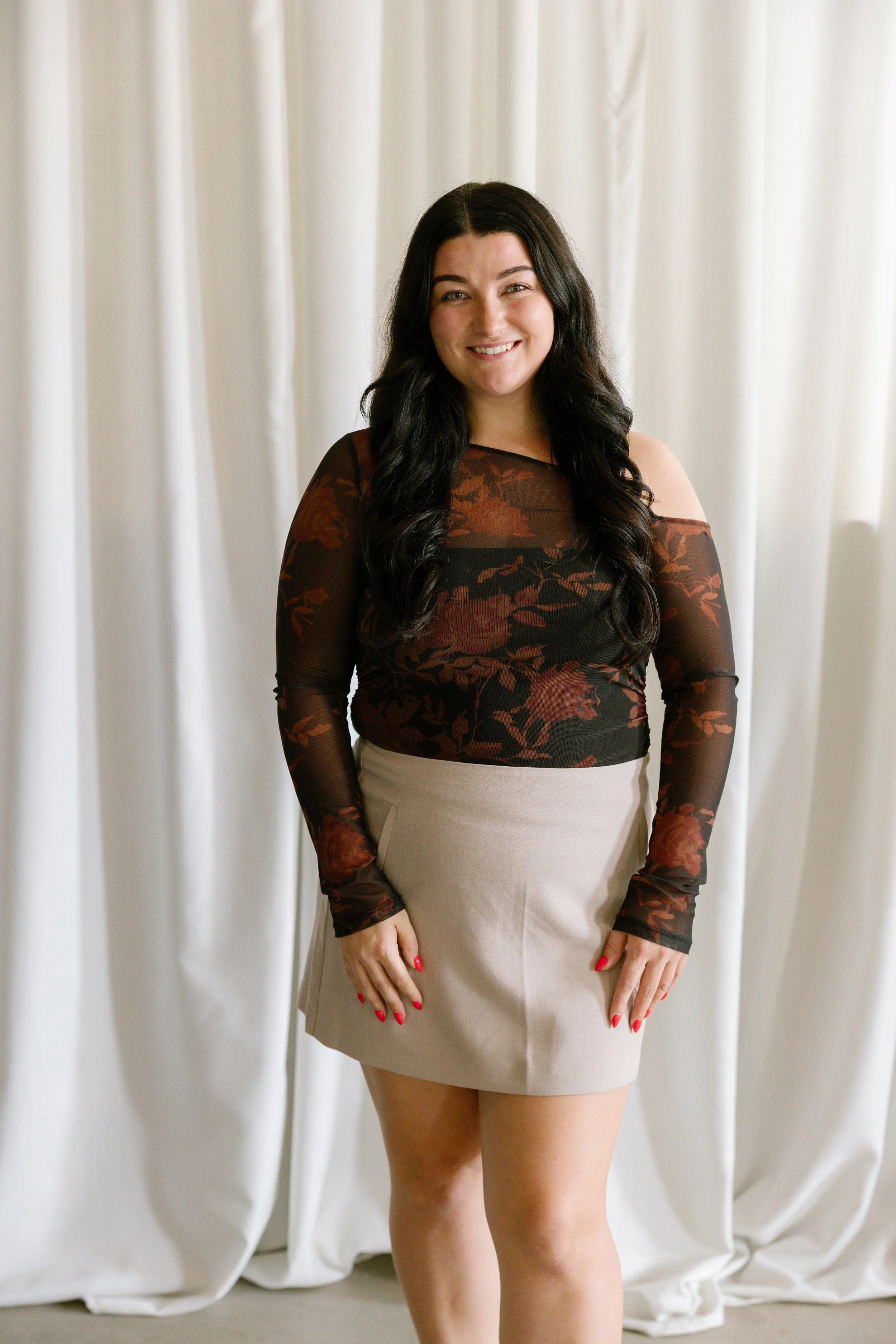 A woman with long dark hair wearing a sheer black top with floral patterns and a beige skirt, standing in front of white curtains, smiling at the camera.