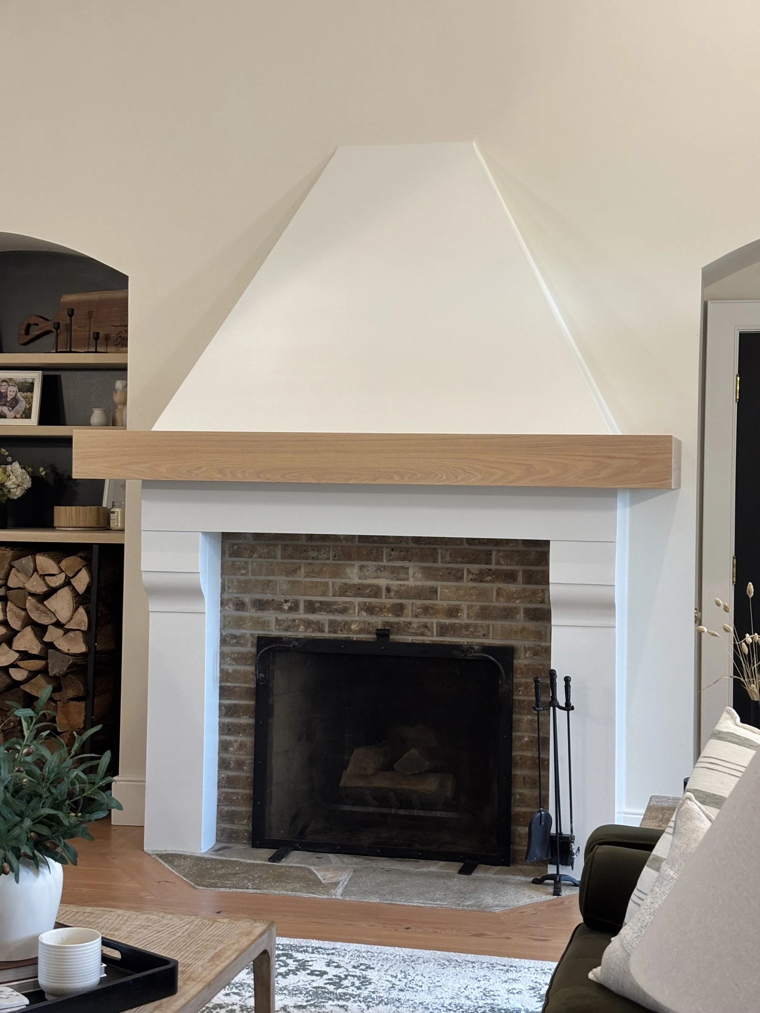 Living room with a fireplace featuring a brick interior and white mantel, flanked by white columns, with a wooden shelf above. To the left, a black bookshelf holds various decorative items and firewood. To the right, a partially visible sofa with pillows, and a side table with a plant and cups.