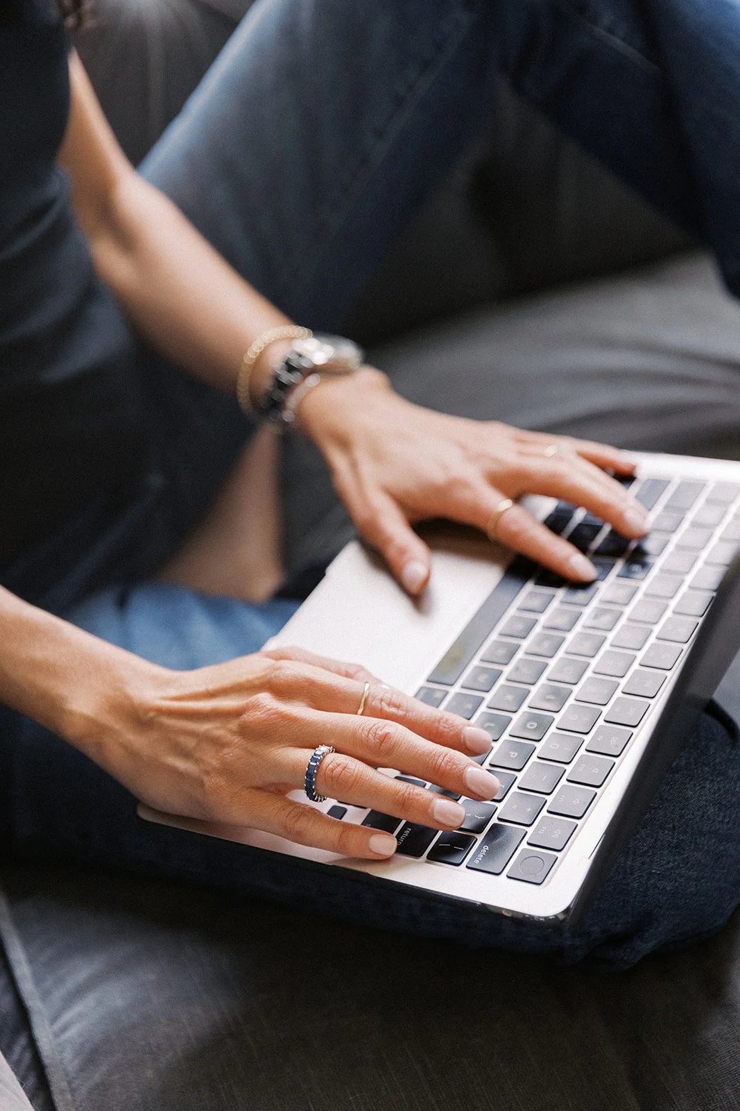 Person using a laptop computer with hands on the keyboard, seated on a dark-colored couch.