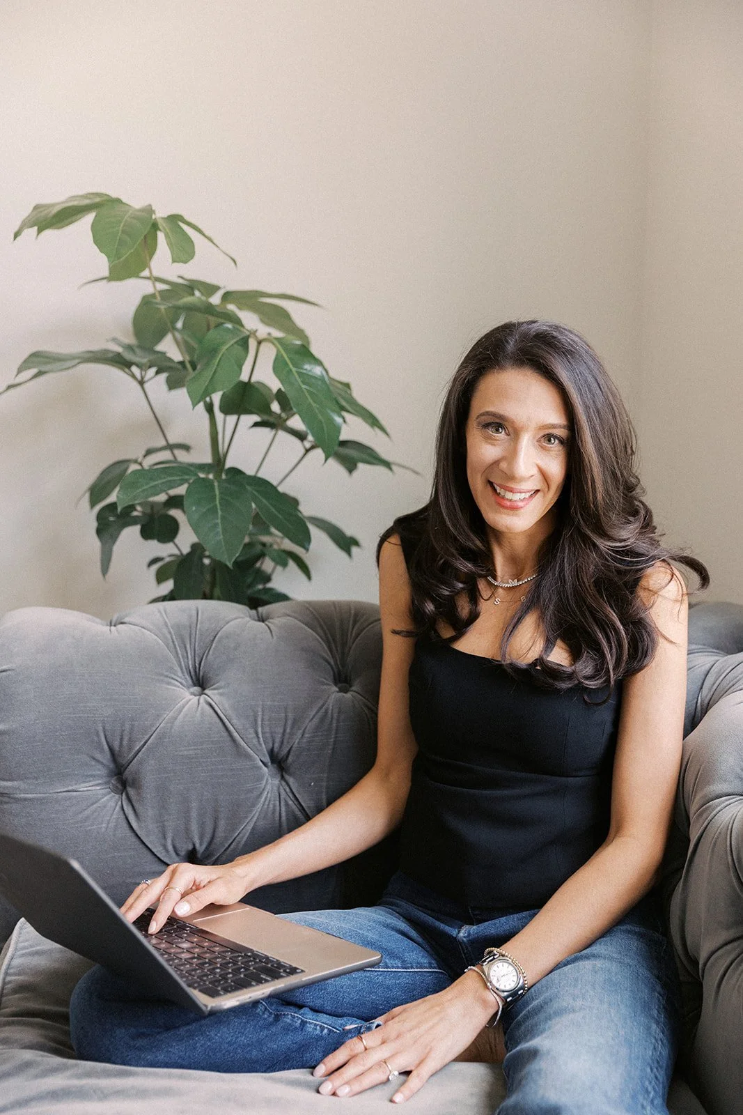 A woman with long dark hair sitting on a gray tufted couch with a laptop on her lap, smiling at the camera. There is a green leafy plant behind her against a plain white wall.