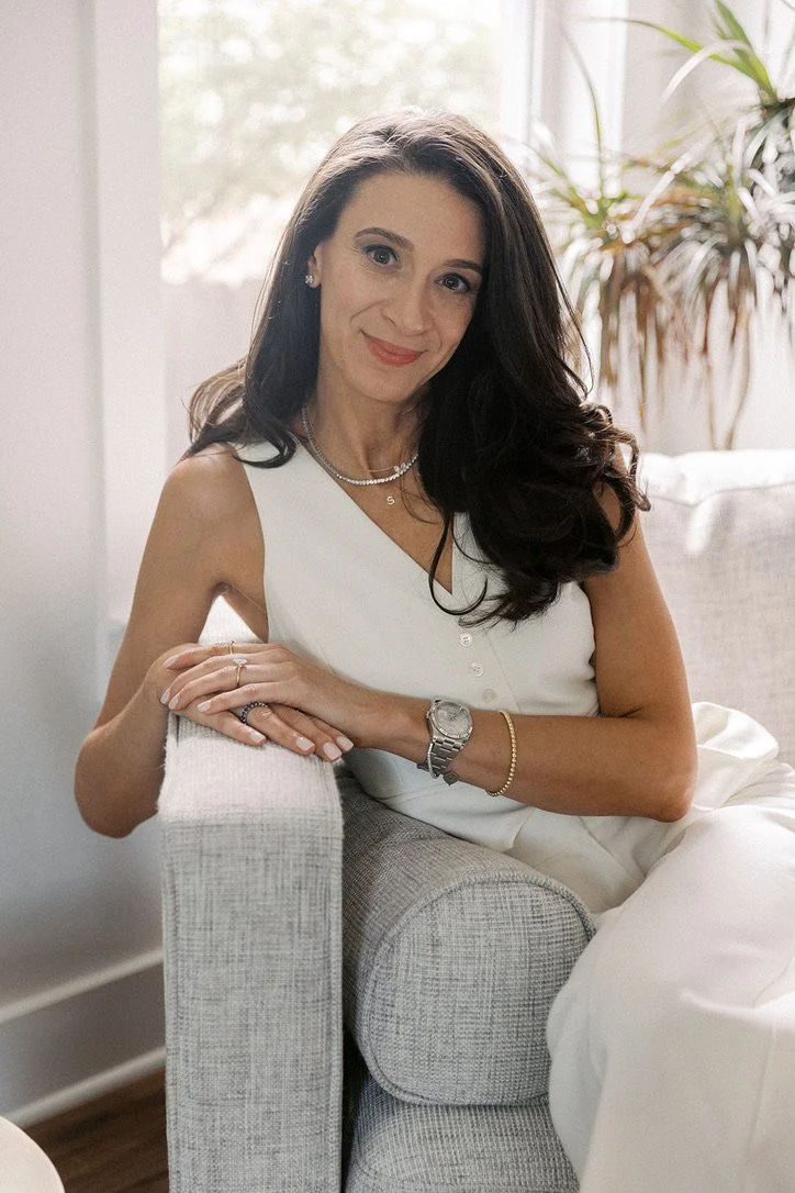 A woman with long dark hair, wearing a white sleeveless top, sitting on a light-colored sofa, smiling and looking at the camera. She has jewelry, including a watch, bracelet, and necklaces, and is in a bright room with sunlight and a potted plant in the background.