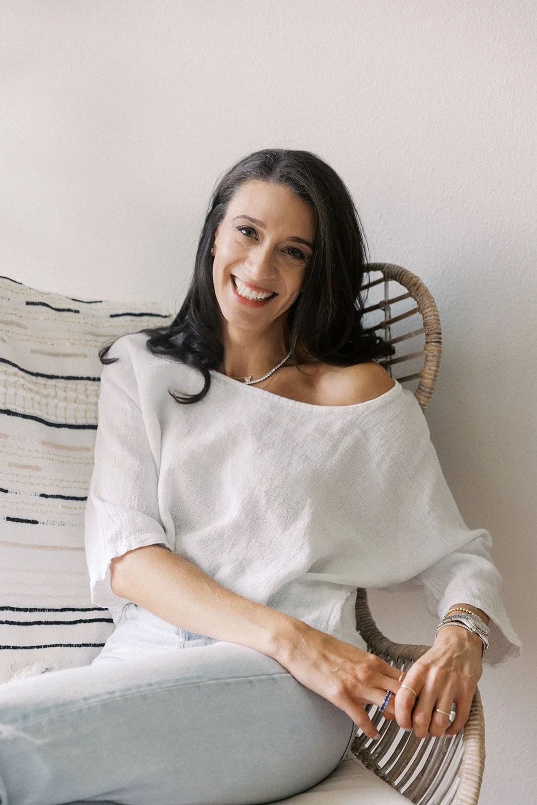 A woman with long dark hair and a bright smile, sitting on a woven chair with a white wall background, wearing a white off-the-shoulder top, multiple rings, bracelets, and necklaces.