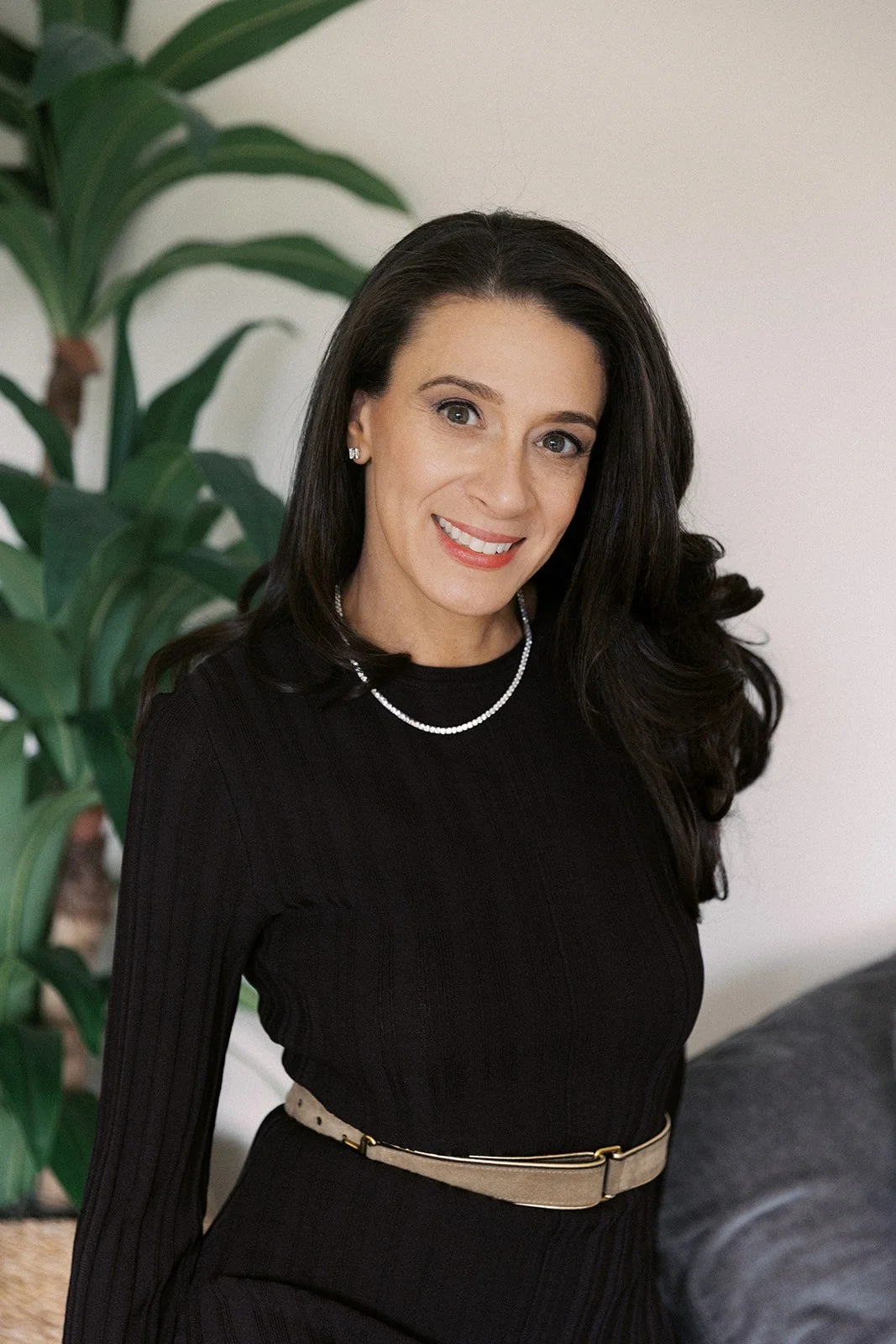 A woman with long dark hair, wearing a black top, a pearl necklace, and earrings, smiling indoors with a green plant and white wall in the background.