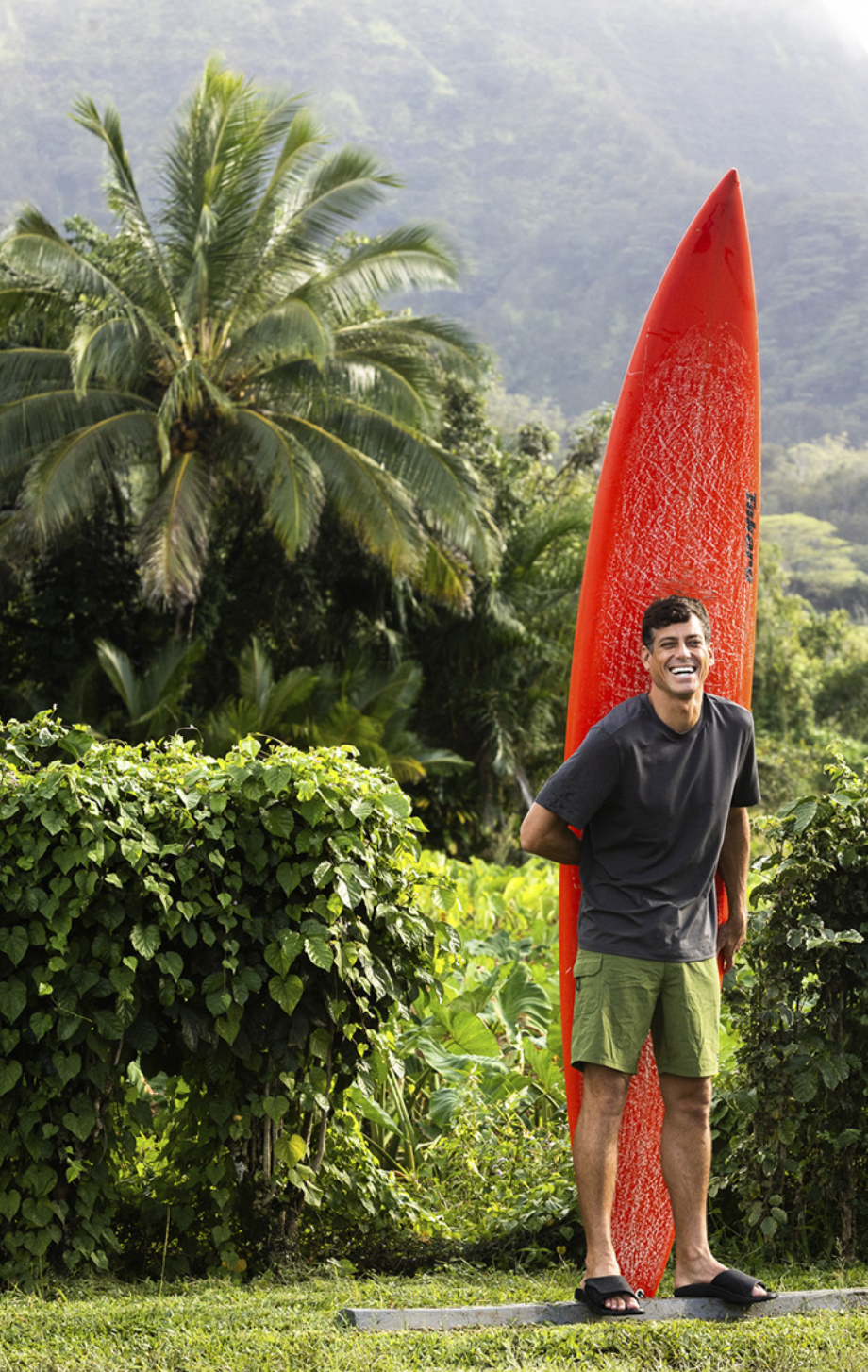 A man in a black t-shirt and green shorts standing next to a red surfboard with a tropical background of palm trees and lush green foliage.
