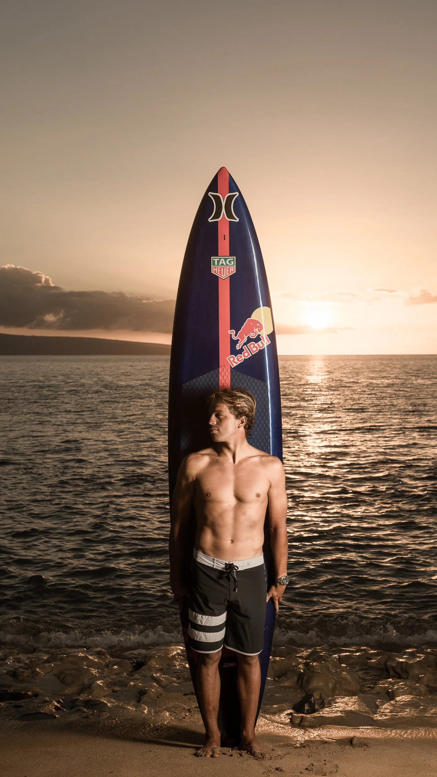 A young man standing on the beach at sunset, shirtless, holding a surfboard with a Red Bull and other logos, with the ocean and sunset in the background.