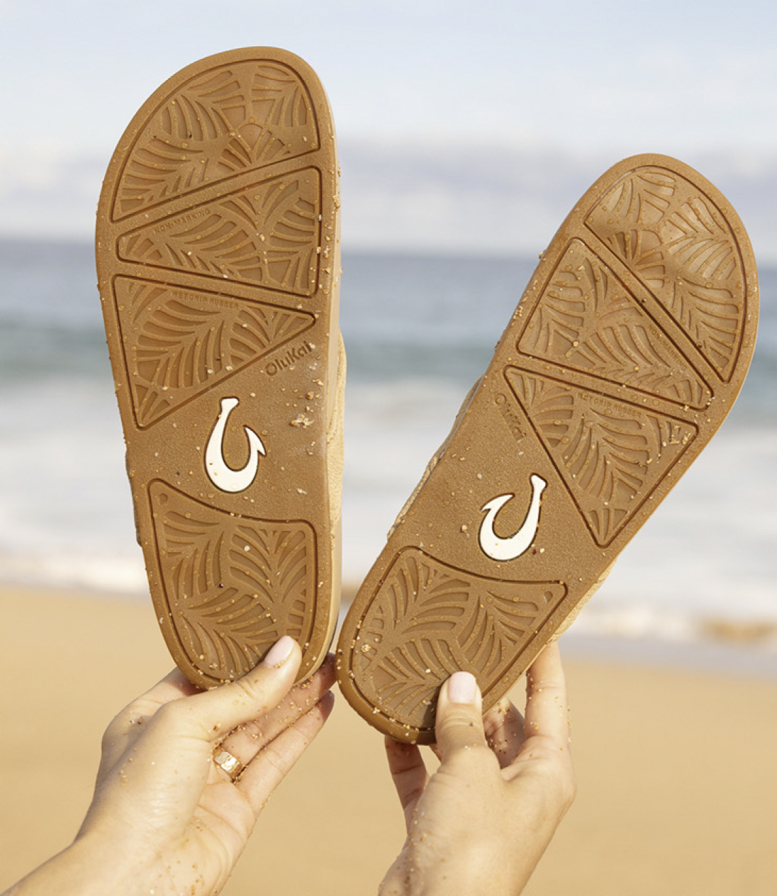 Two hands holding brown flip-flops with leaf patterns and a white horseshoe logo, against a beach and ocean background.