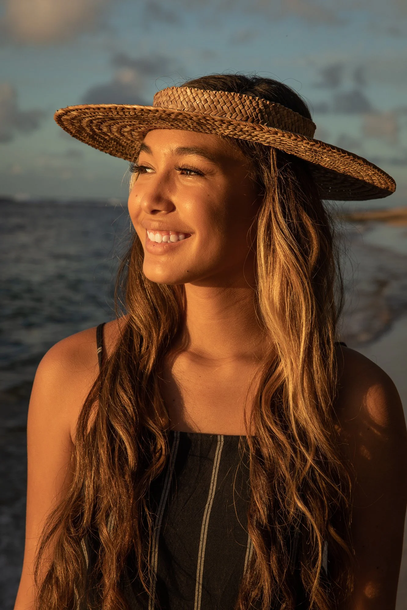 A woman with long wavy hair wearing a wide-brimmed straw hat, smiling and looking into the distance at sunset by the ocean.