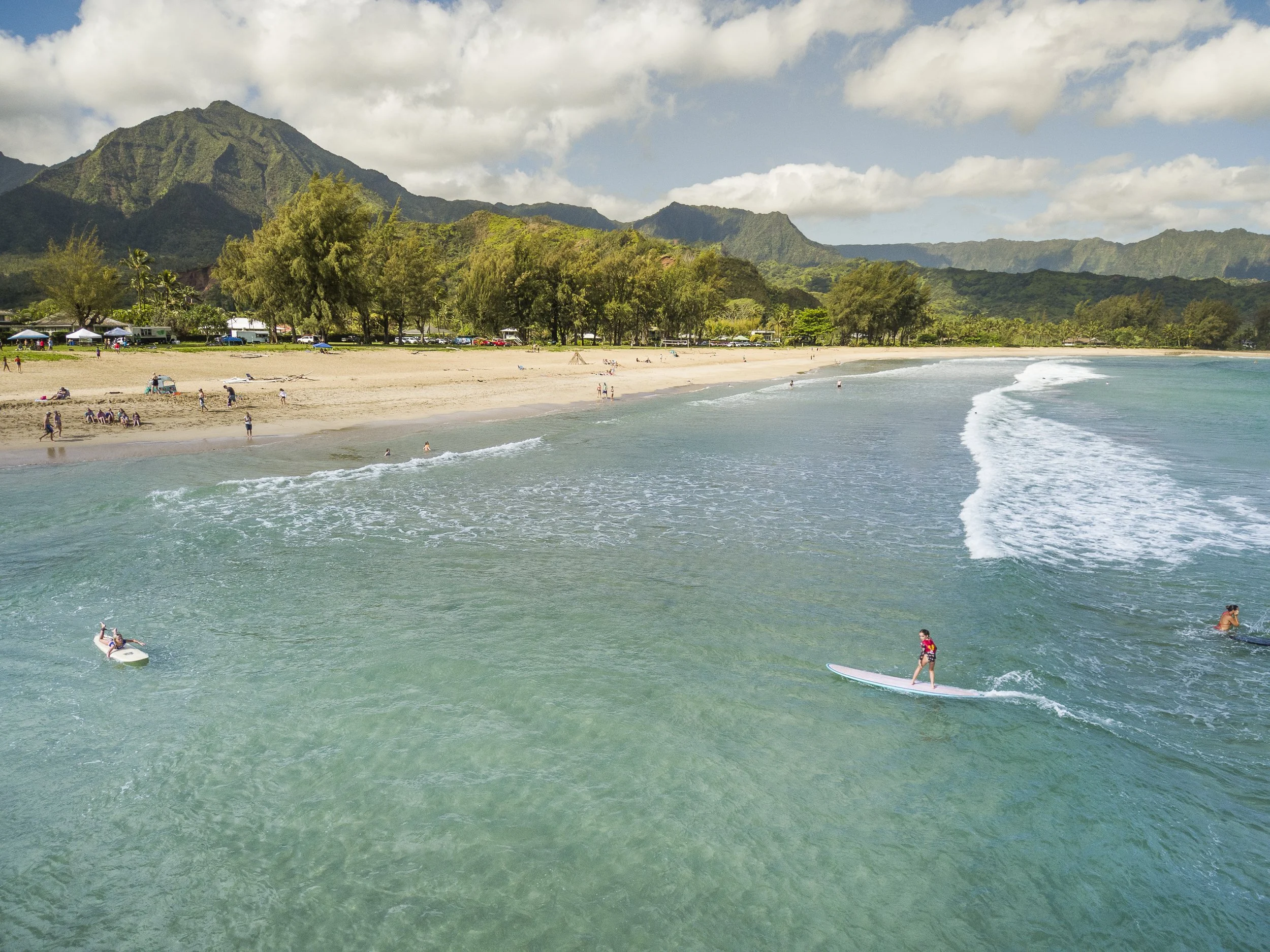 A scenic beach with a mountain backdrop, people swimming and surfing in the ocean, and umbrellas and trees along the sandy shore.