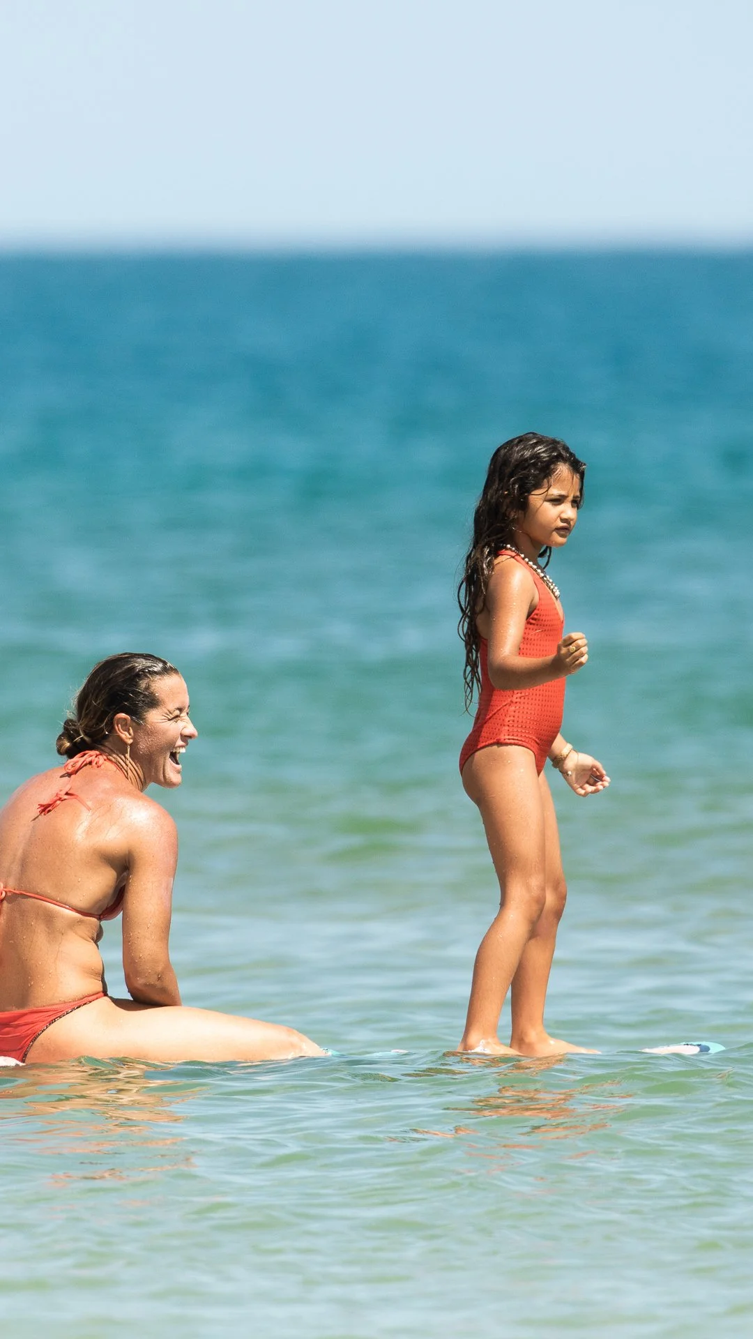 A woman and a girl in red swimsuits enjoying time at the beach, with the woman laughing and the girl standing on a surfboard in the shallow water.