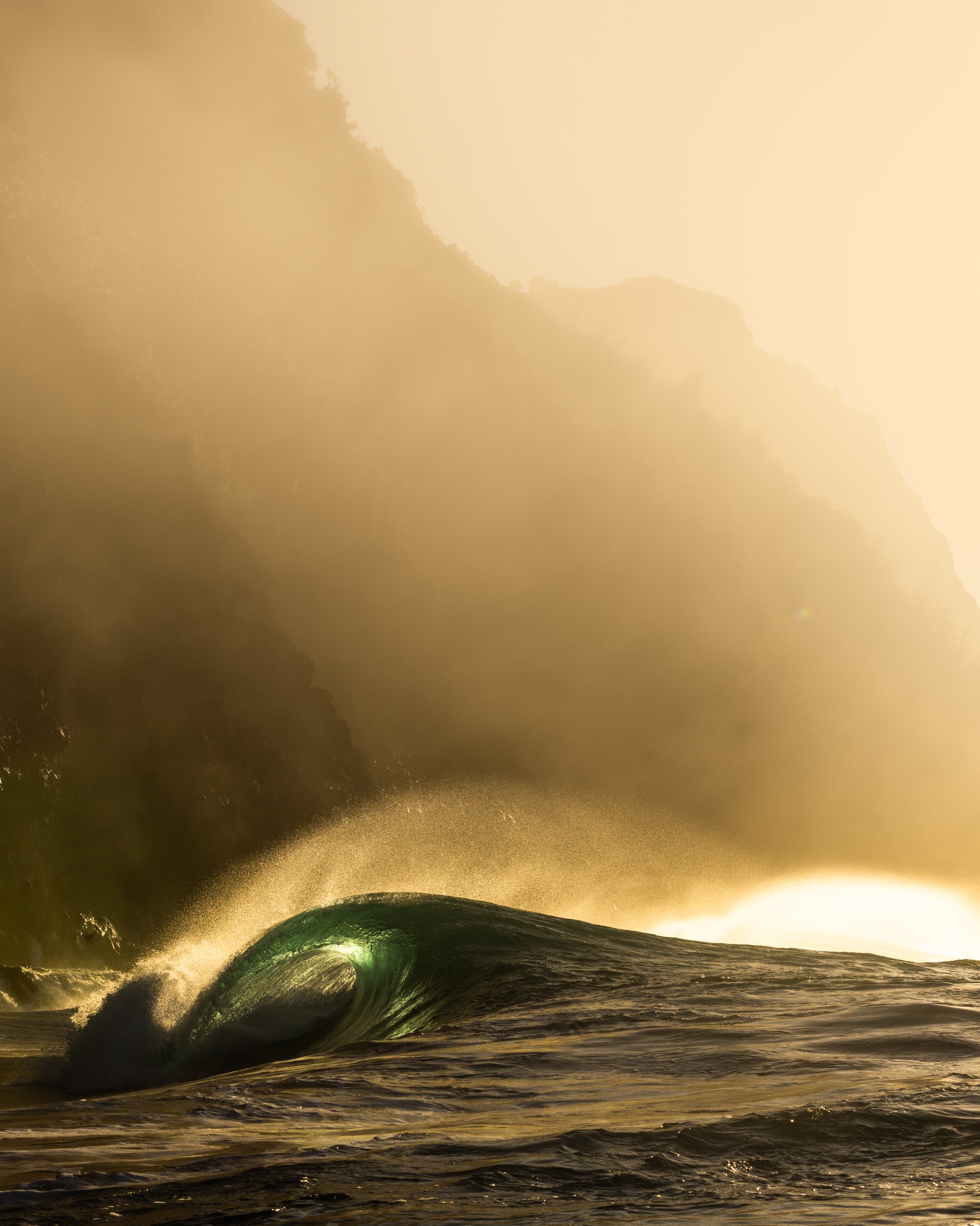 A large ocean wave crashing with spray and mist, illuminated by golden sunlight, with a shoreline and hills in the background.